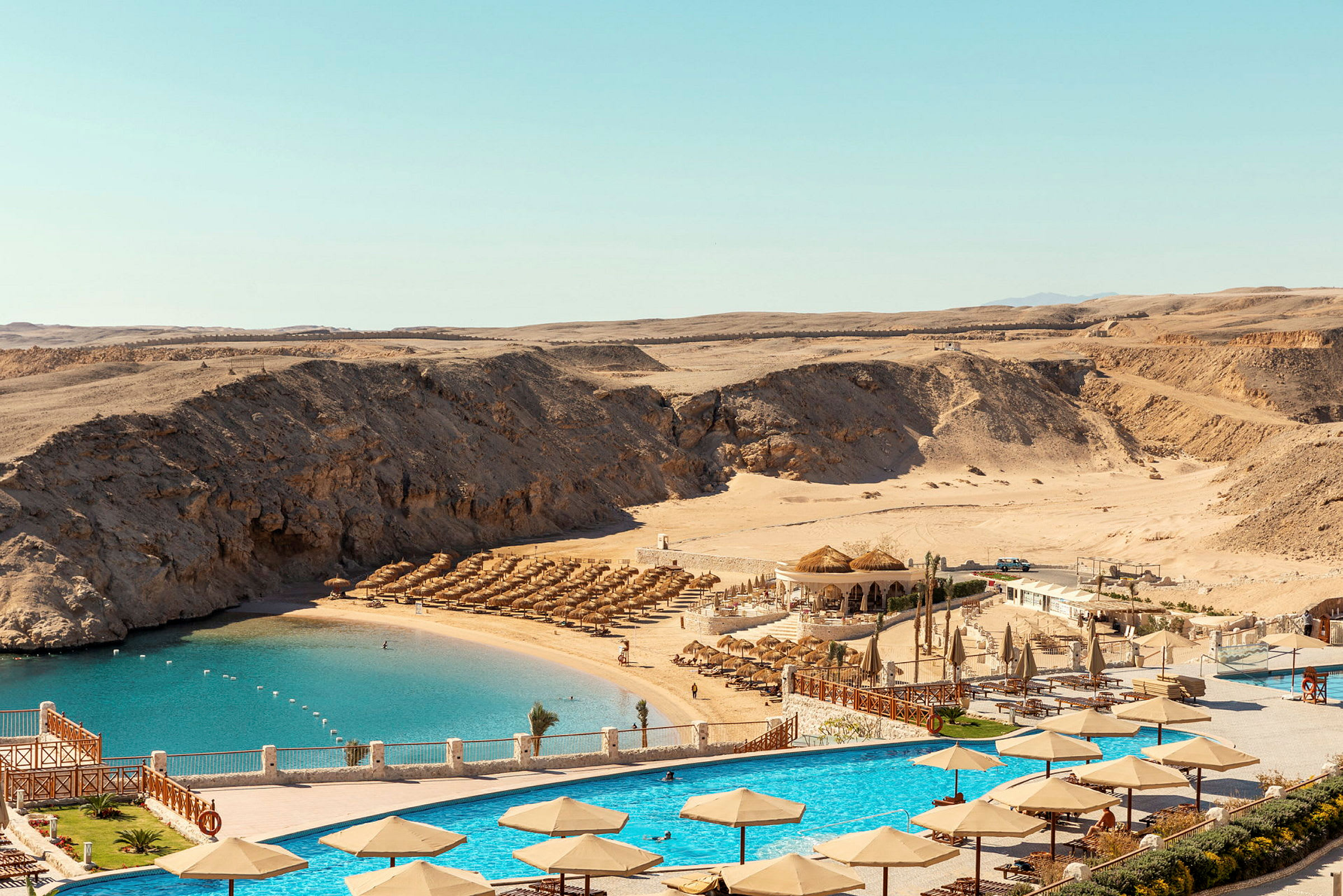 Vy över en resort med pool och parasoller, strand med solstolar och kringliggande sanddyner. (AI-genererad text)