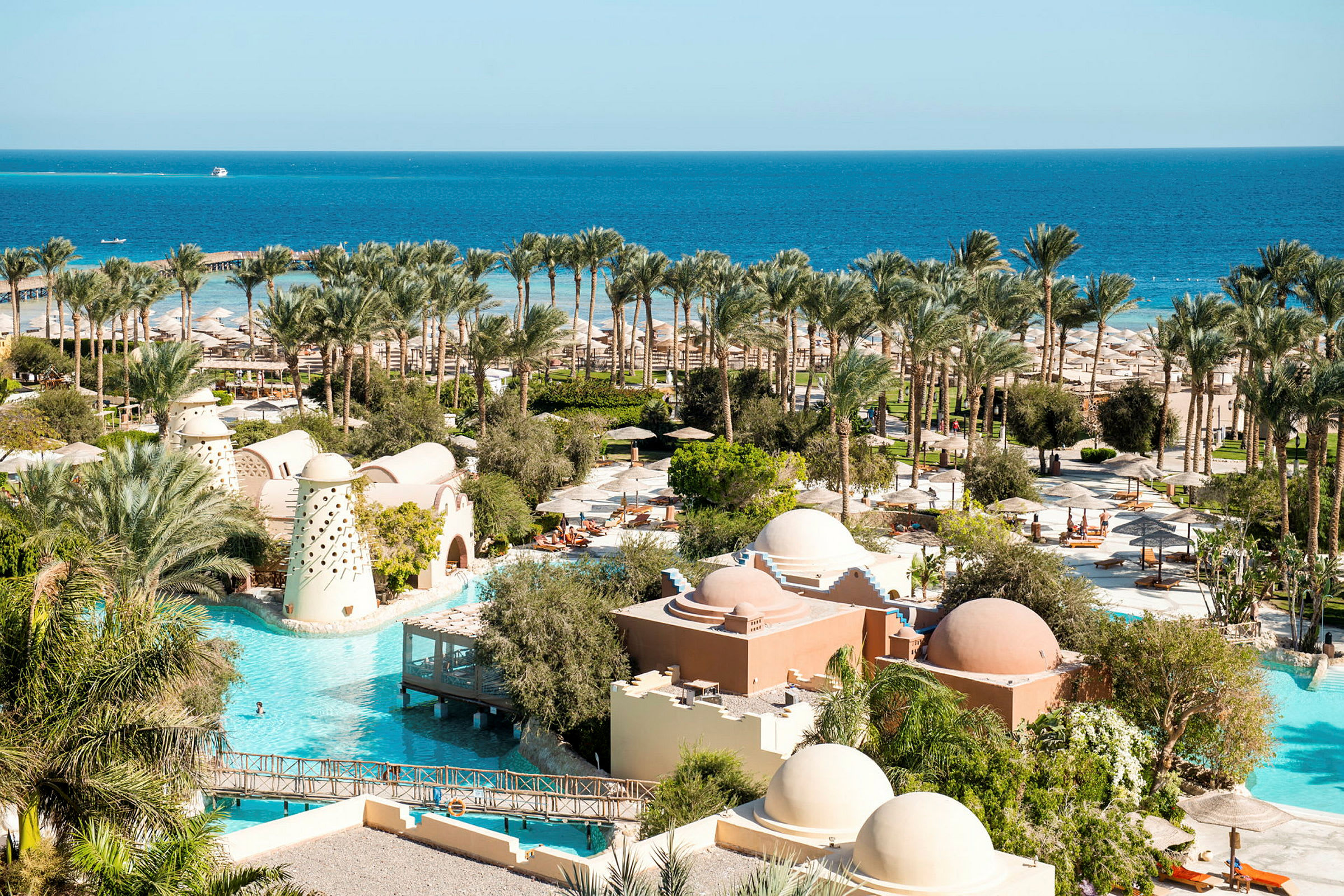 Bild av ett hotellområde med pooler, palmer och byggnader med kupoler, i närheten av en kust med havsutsikt. (AI-genererad text)