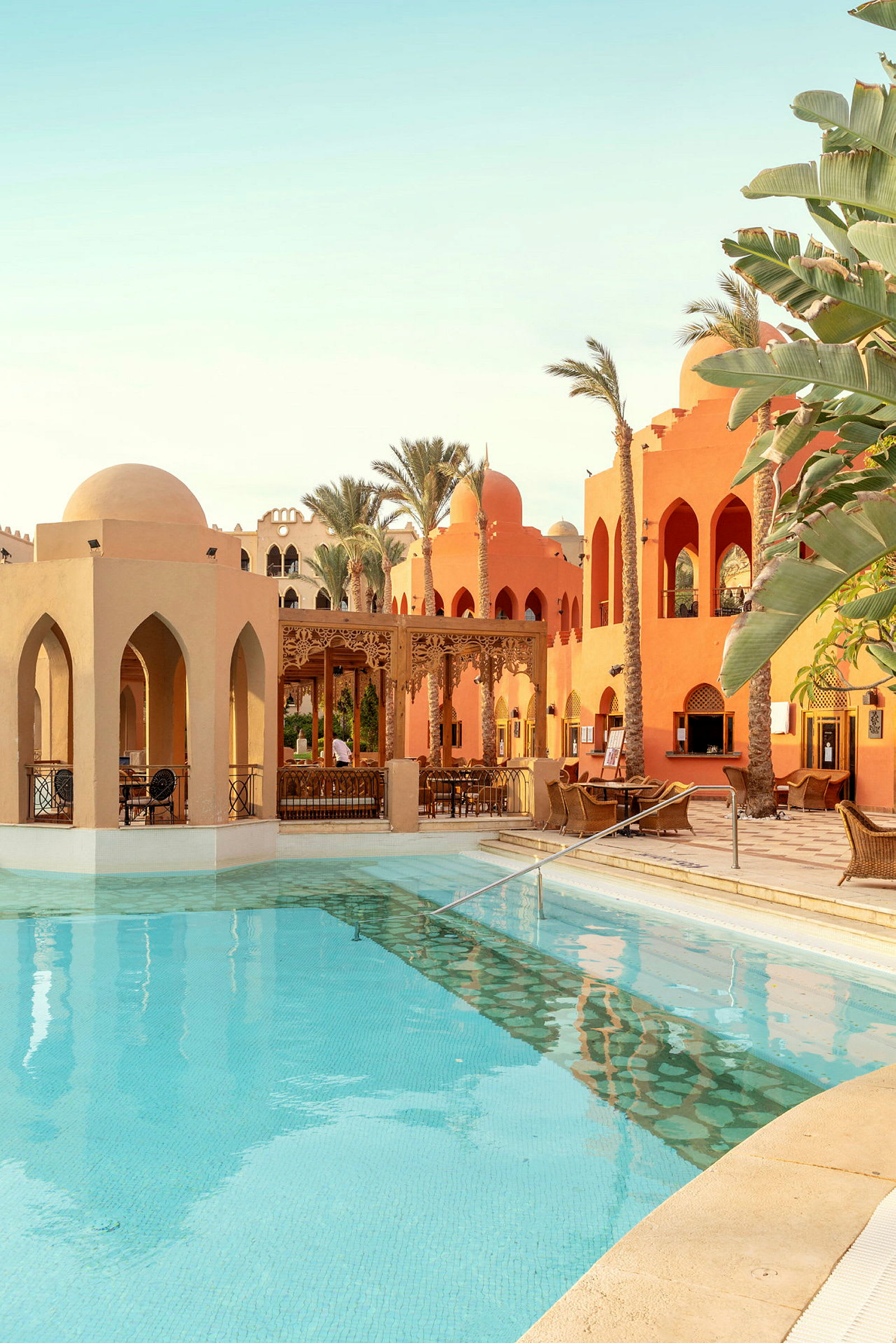En pool med klart vatten omgiven av byggnader med arabisk arkitektur och palmträd. (AI-genererad text)