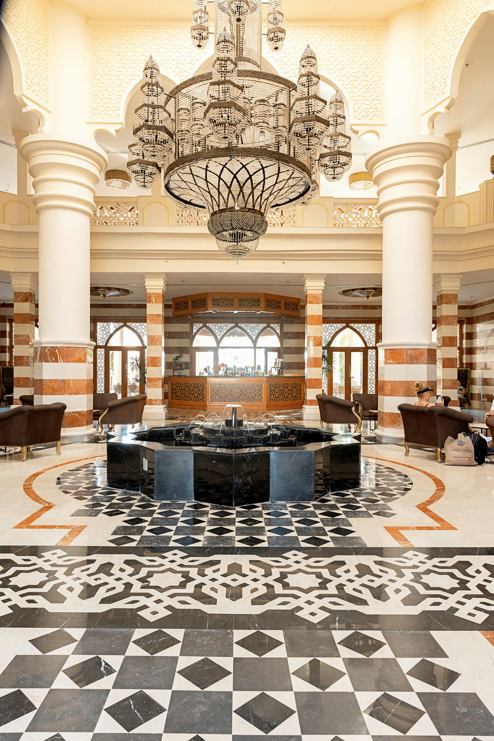 Elegant hotellobby med mönstrad golvbeläggning, fontän i mitten och en stor kristallkrona hängande ovanför. (AI-genererad text)