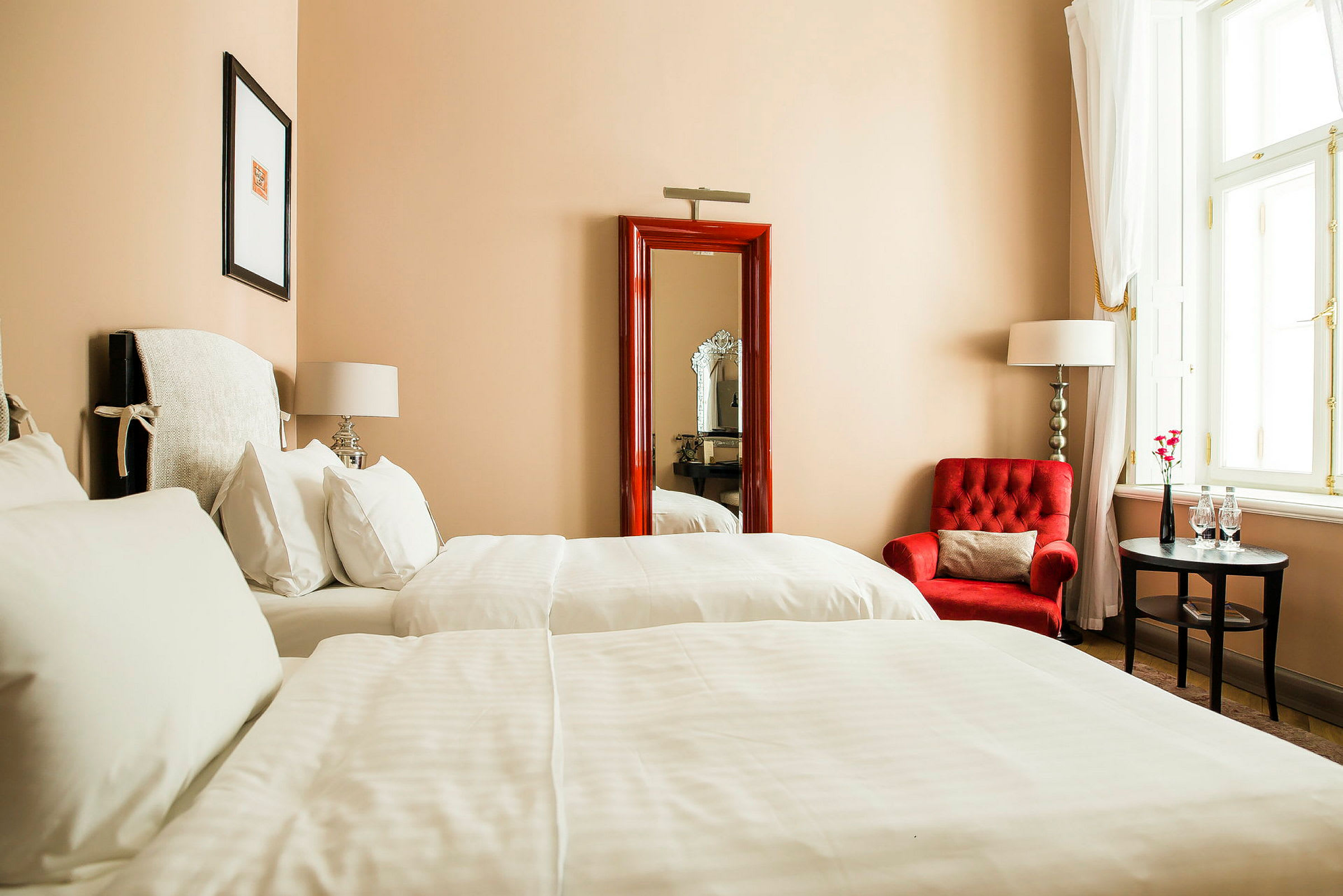 Ett hotellrum med två bäddar, en röd fåtölj, ett litet bord och ett stort fönster med ljusa gardiner. (AI-genererad text)