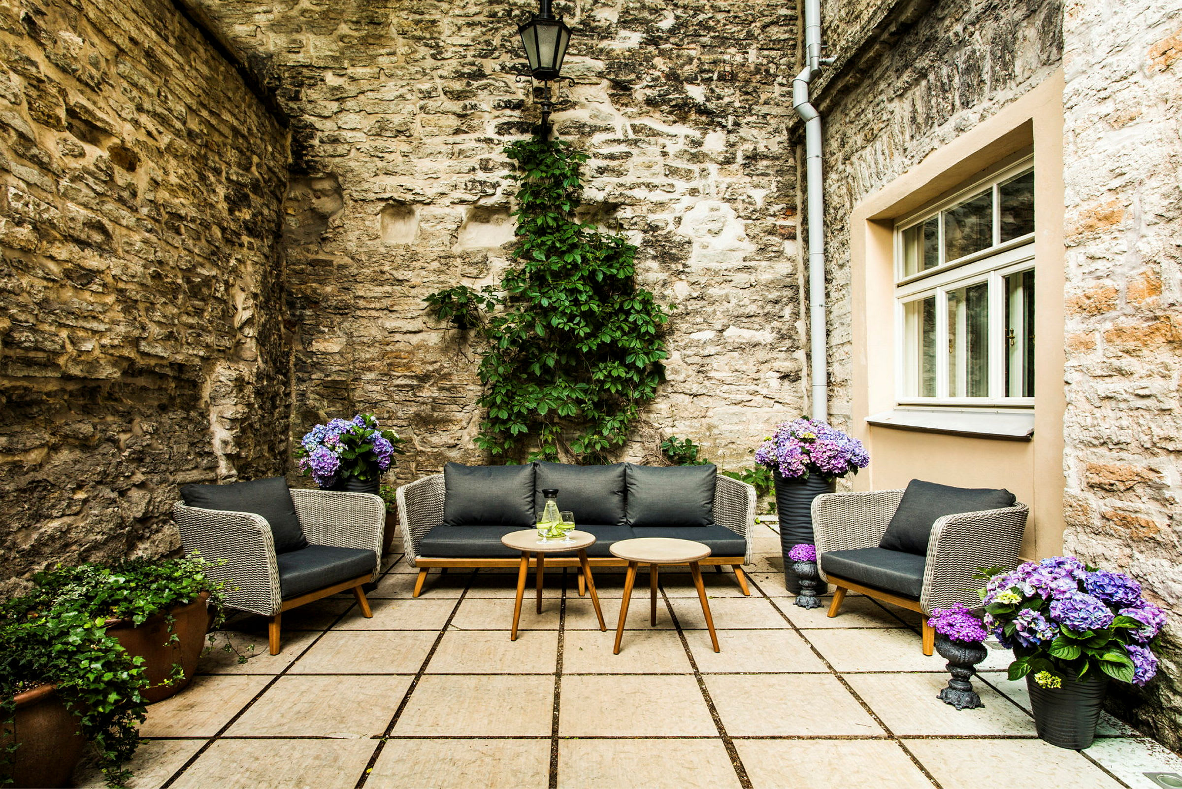 En innergård med stenlagd mark, stenväggar och mysiga sittplatser med kuddar. Krukor med blommor och grönska dekorerar området. (AI-genererad text)