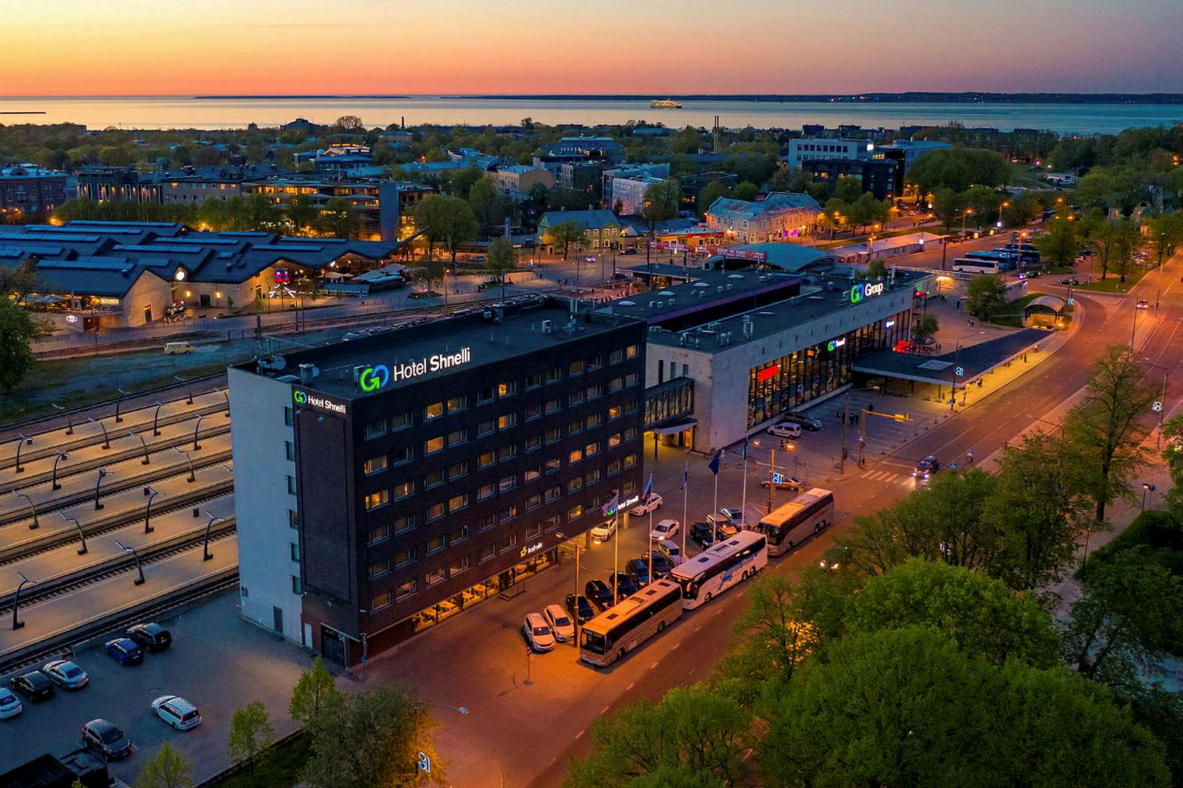 En större byggnad med skylten "Hotel Shnelli" syns vid en väg omgiven av bussar och bilar. Området är upplyst på kvällen med en bakgrund av träd och en hamn. (AI-genererad text)