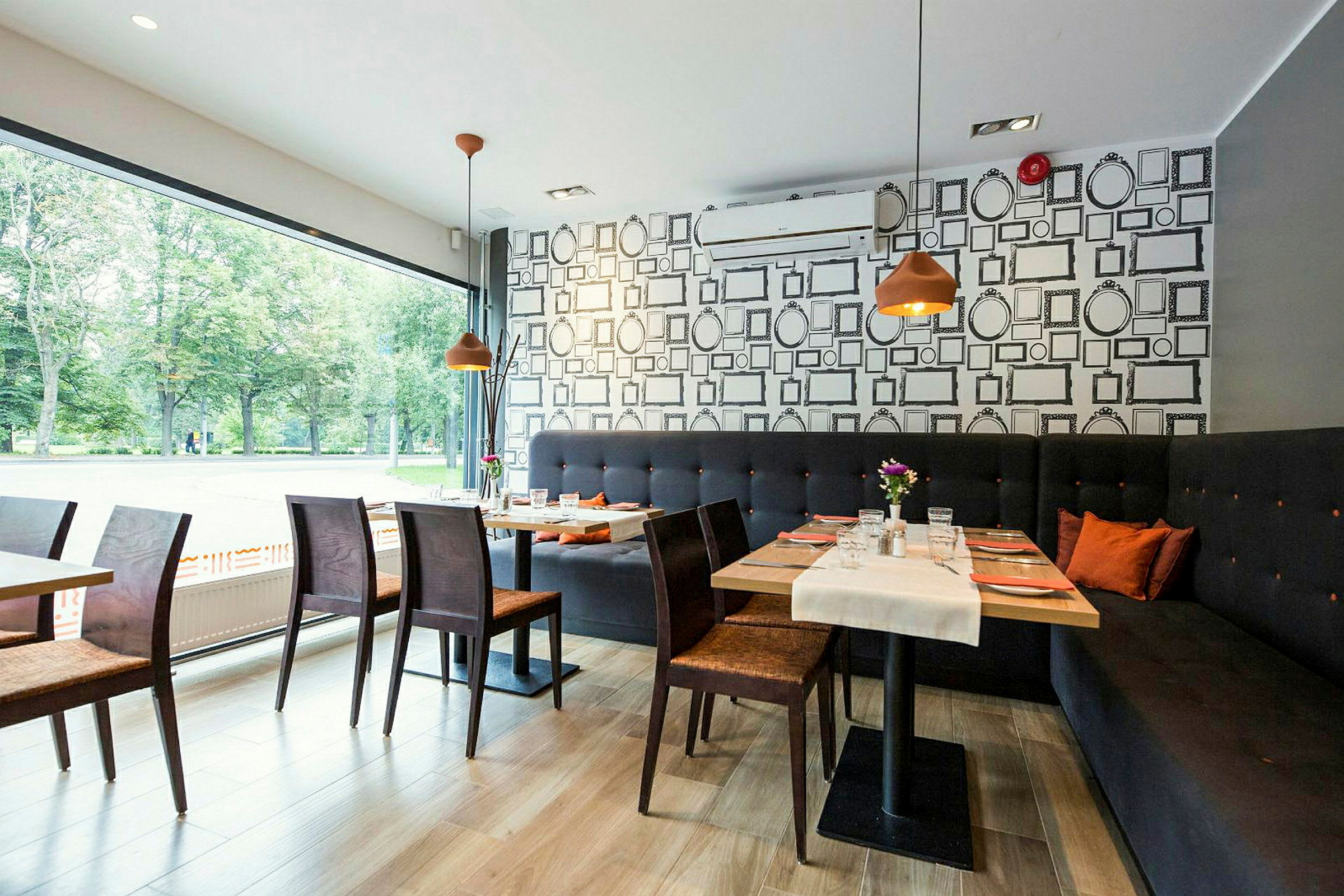 Ett modernt inrett kafé eller restaurang med stora fönster, träbord och stolar samt en lång soffa längs väggen. (AI-genererad text)