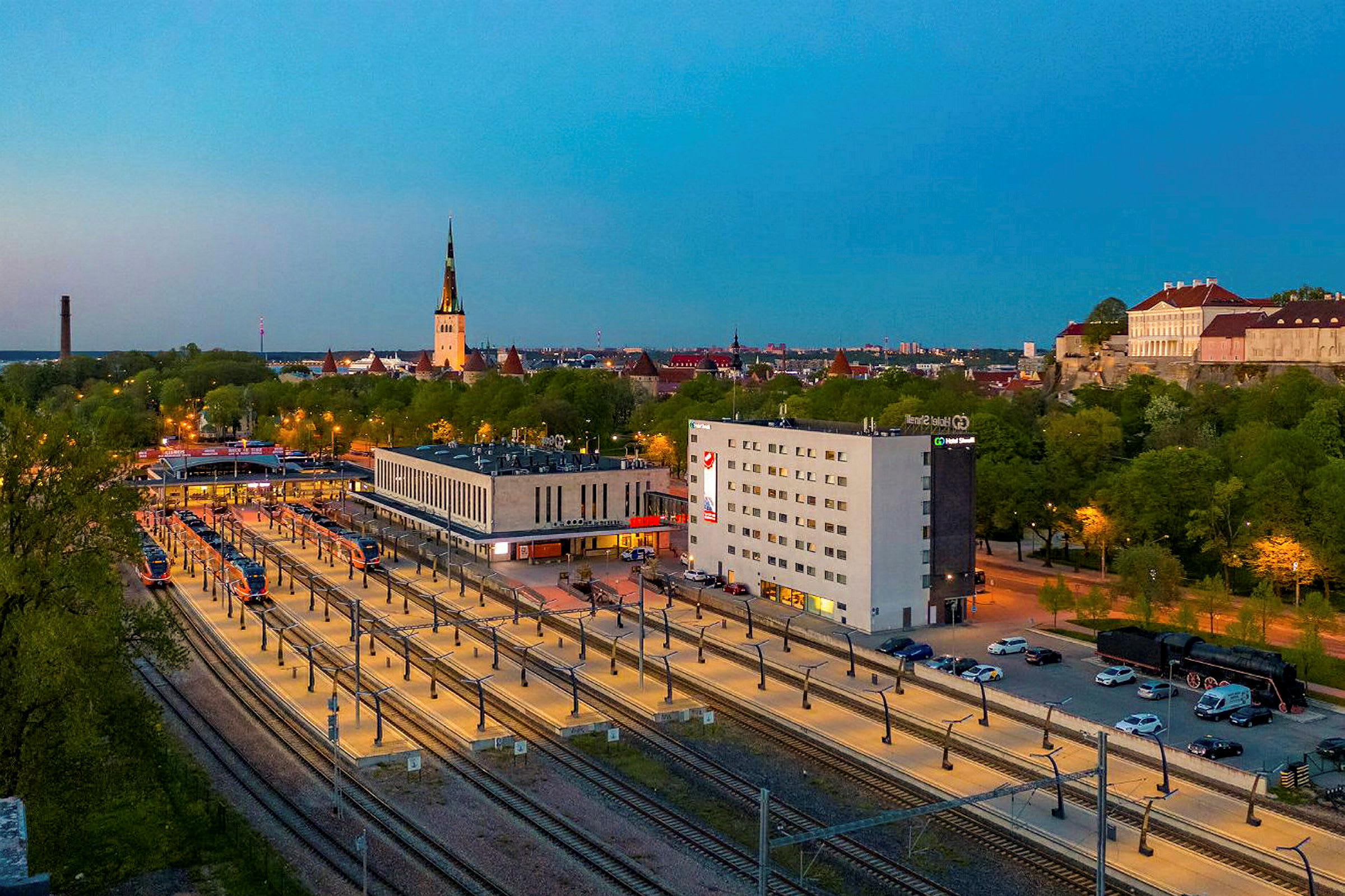 Bild av en tågstation omgiven av byggnader och gröna träd, med en kyrkspira synlig i bakgrunden. (AI-genererad text)