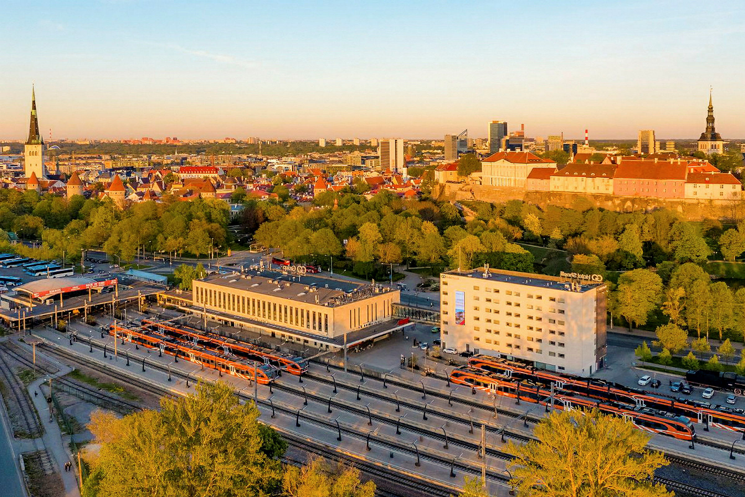 Bild av en stad med järnvägsstation i förgrunden och stadens silhuett i bakgrunden med byggnader och träd. (AI-genererad text)
