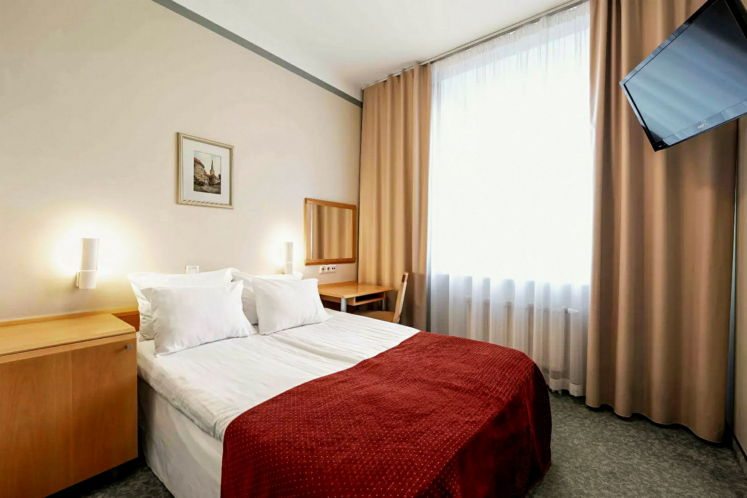 Ett hotellrum med en dubbelsäng, rödfärgad sängöverkast. En spegel och skrivbord står intill väggen. Ett fönster med ljusa gardiner och en väggmonterad TV finns i rummet. (AI-genererad text)