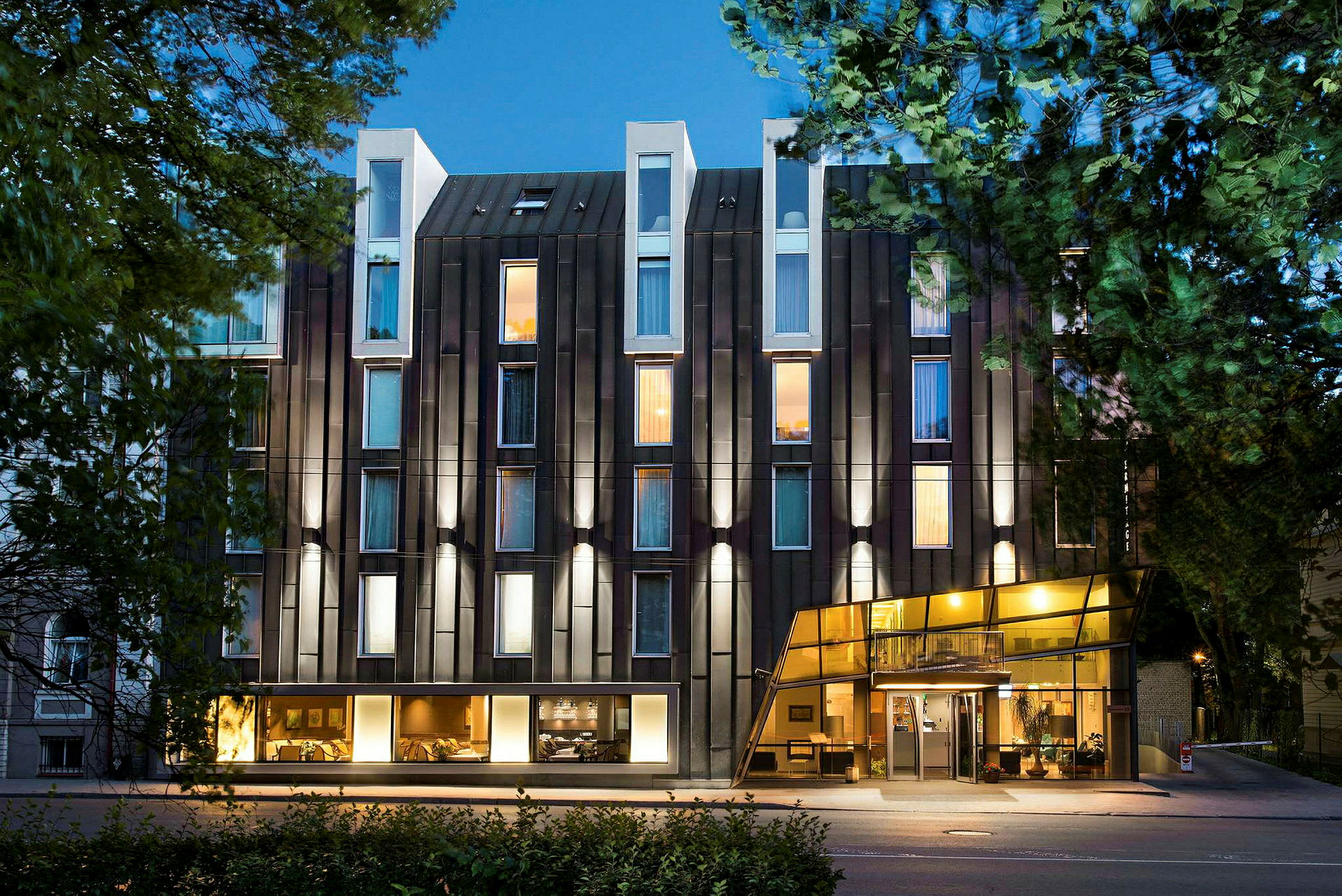 Exteriör av ett modernt hotell med glasfasad och upplysta fönster omgiven av träd. (AI-genererad text)