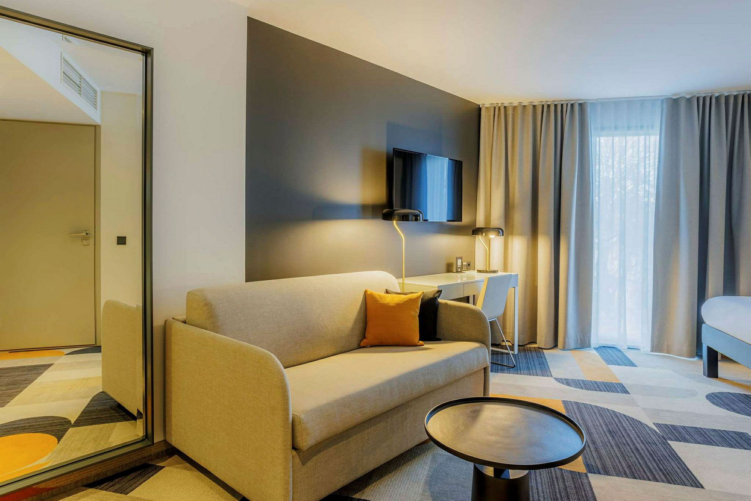 Modernt hotellrum med beige soffa, svart bord, skrivbord med stol och TV på väggen. (AI-genererad text)