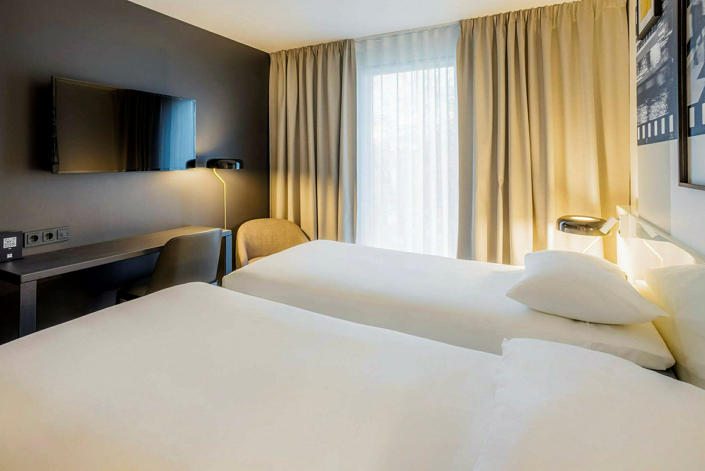 Hotellrum med två enkelsängar, ett skrivbord, en stol och en väggmonterad TV, samt gardiner vid ett fönster. (AI-genererad text)