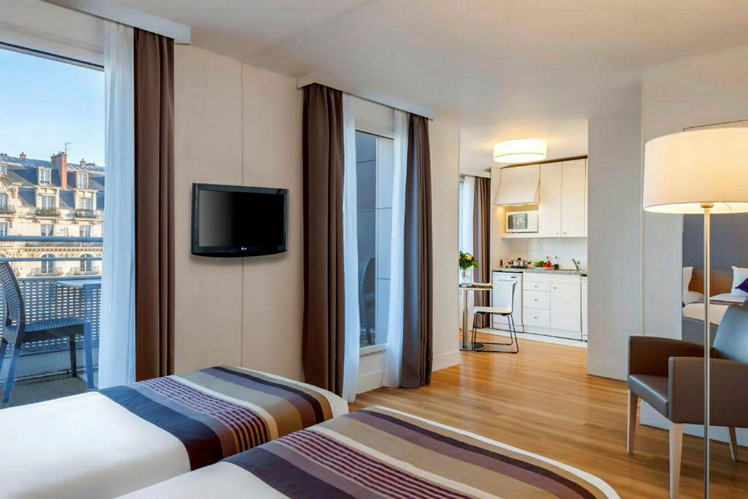 Hotellrum med två sängar, en TV på väggen, ett kök i bakgrunden och en balkong med utsikt över byggnader. (AI-genererad text)
