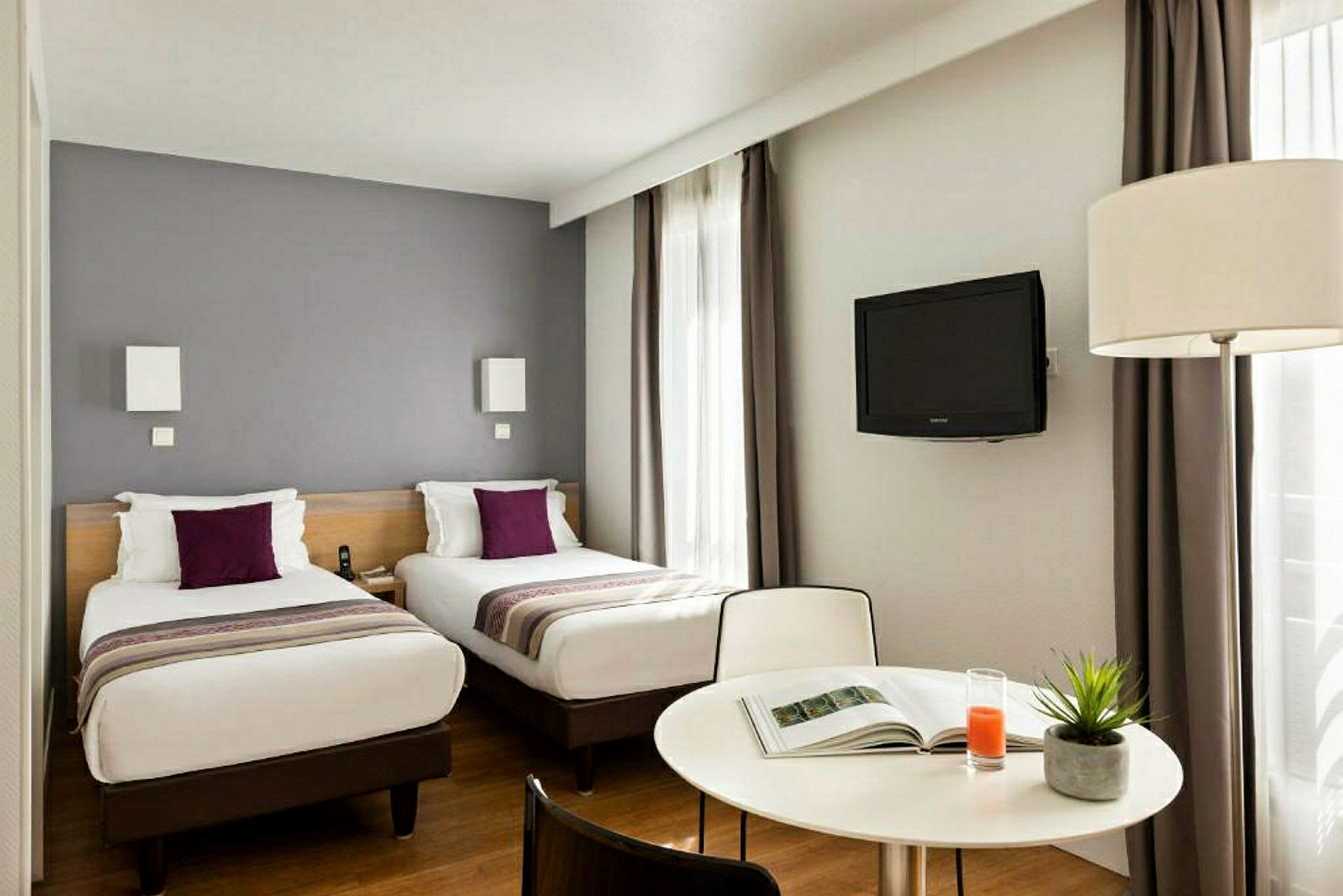 Två enkelsängar med lila kuddar i ett hotellrum, med tv och bord. (AI-genererad text)