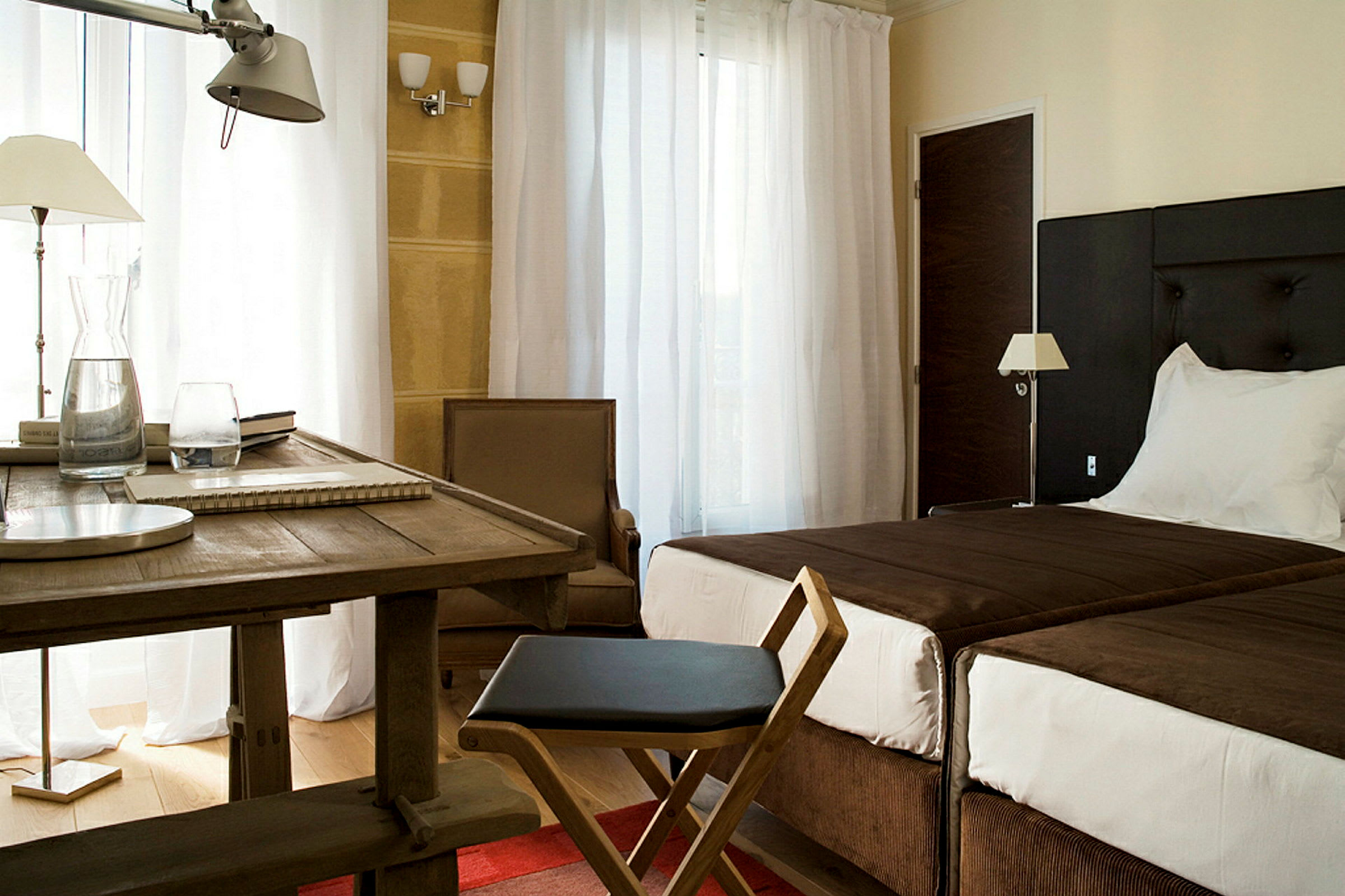 Ett hotellrum med två sängar, ett bord och stol, samt en fåtölj framför vita gardiner. (AI-genererad text)