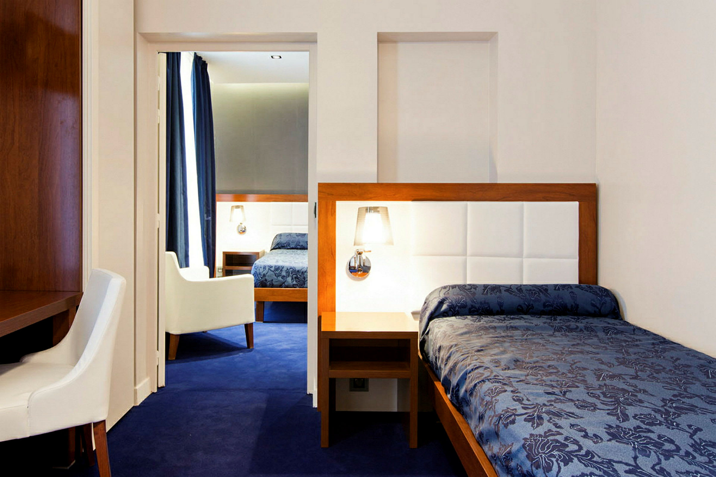 Ett hotellrum med blå matta, en enkelsäng med blått överkast och ljusa möbler. (AI-genererad text)