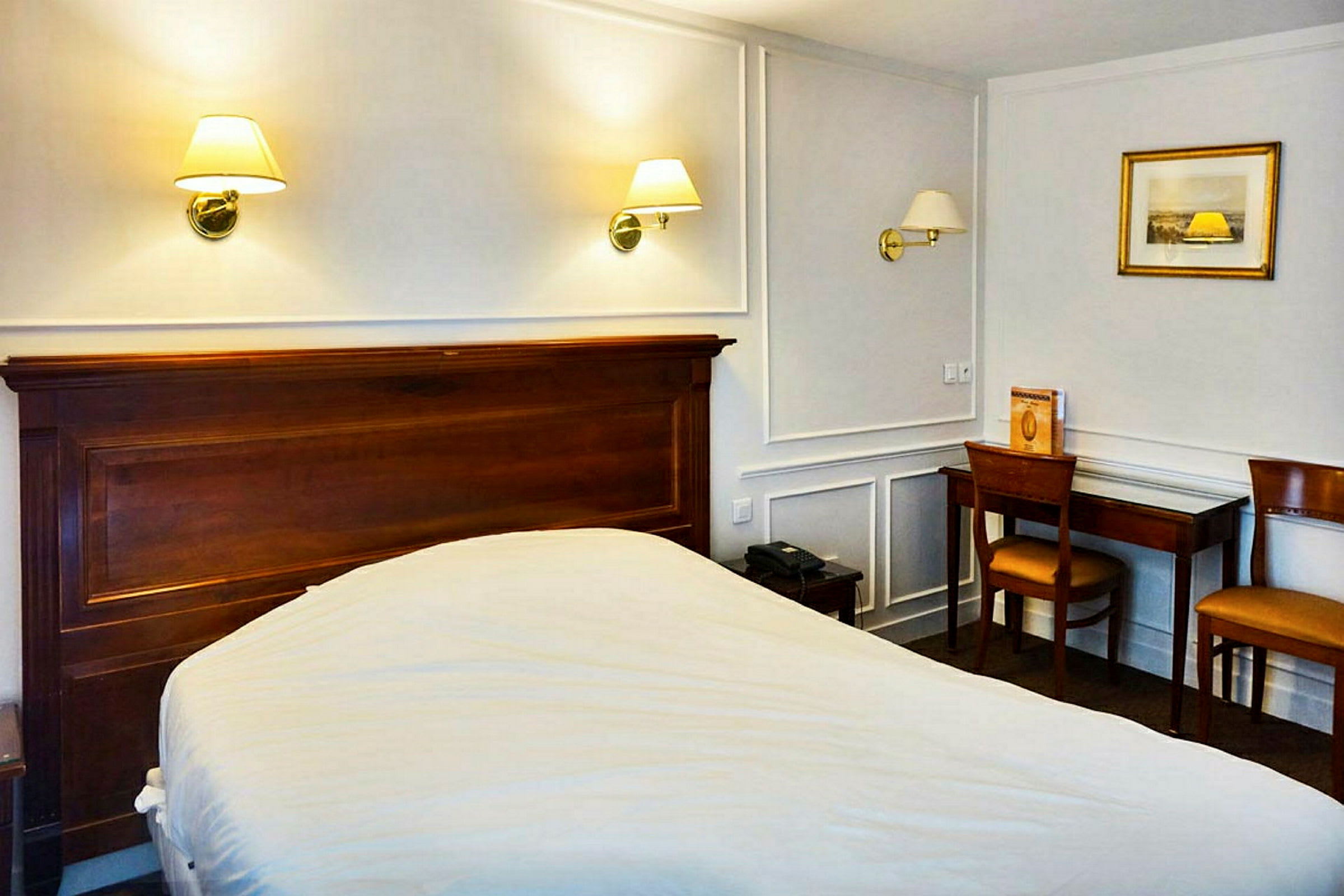 Ett hotellrum med en säng, träbord och stolar samt vägglampor ovanför sängen. (AI-genererad text)