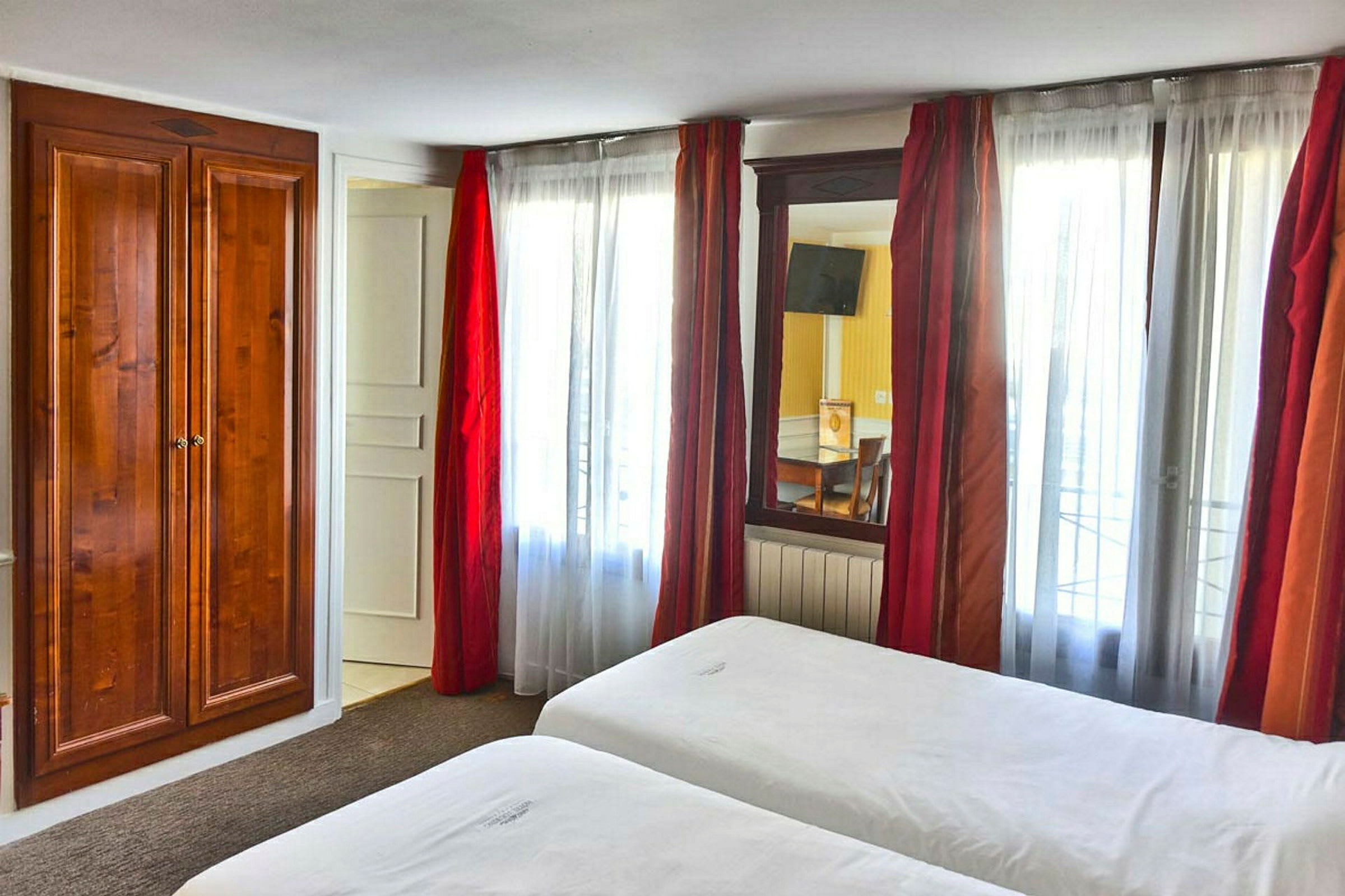 Ett hotellrum med två sängar, vita lakan och en trägarderob bredvid ett fönster med röda gardiner. (AI-genererad text)