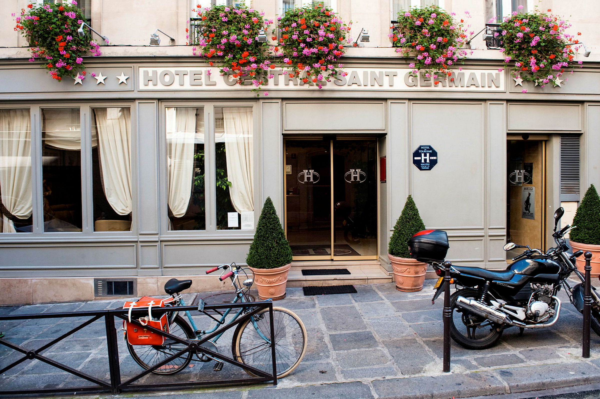 Exteriören av ett hotell med skylten 'Hotel Central Saint Germain' med blomkrukor ovanför ingången samt en cykel och motorcykel parkerad framför. (AI-genererad text)