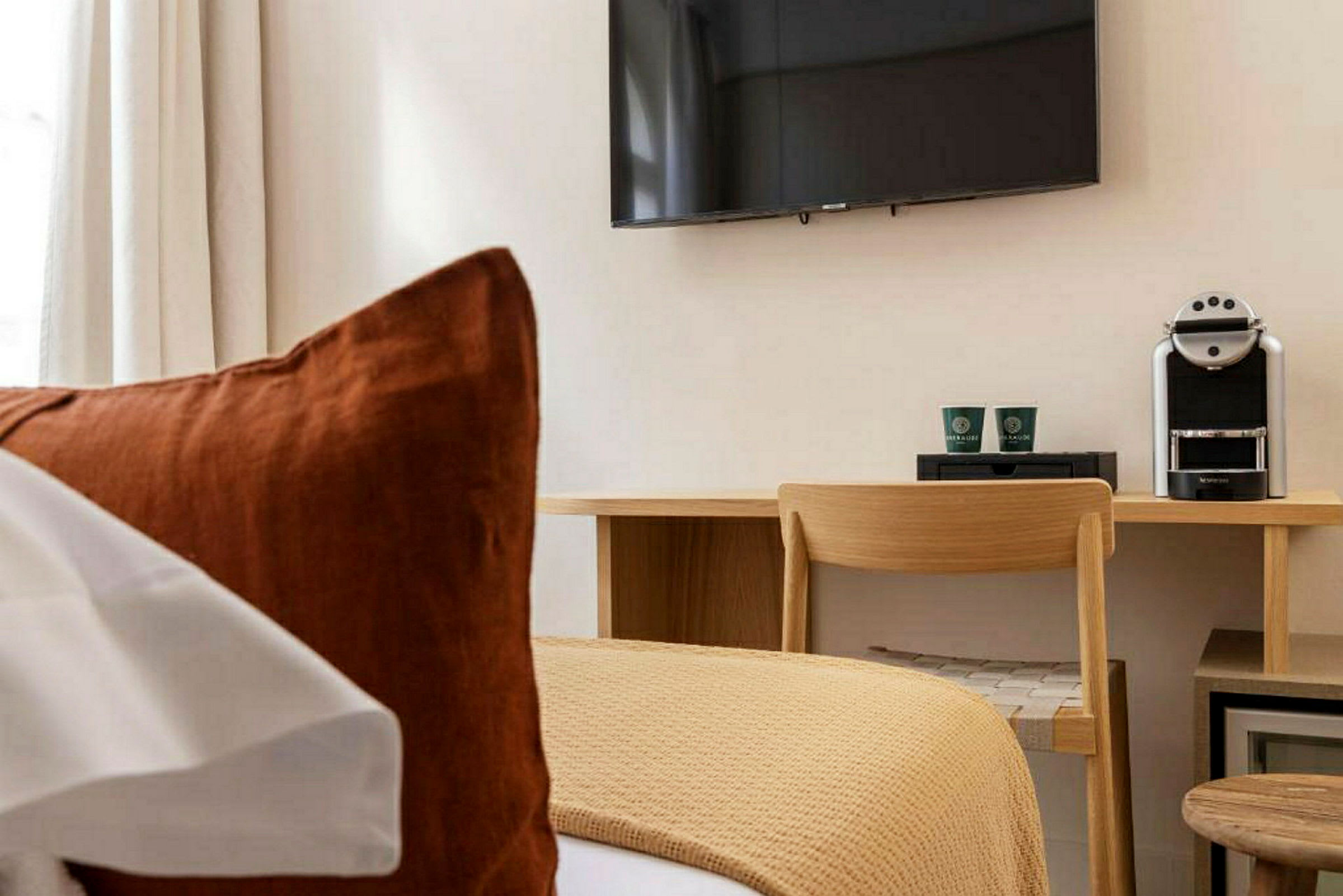 Ett hotellrum med en säng i förgrunden, en skrivbordsplats med stol, kaffemaskin och en väggmonterad TV. (AI-genererad text)