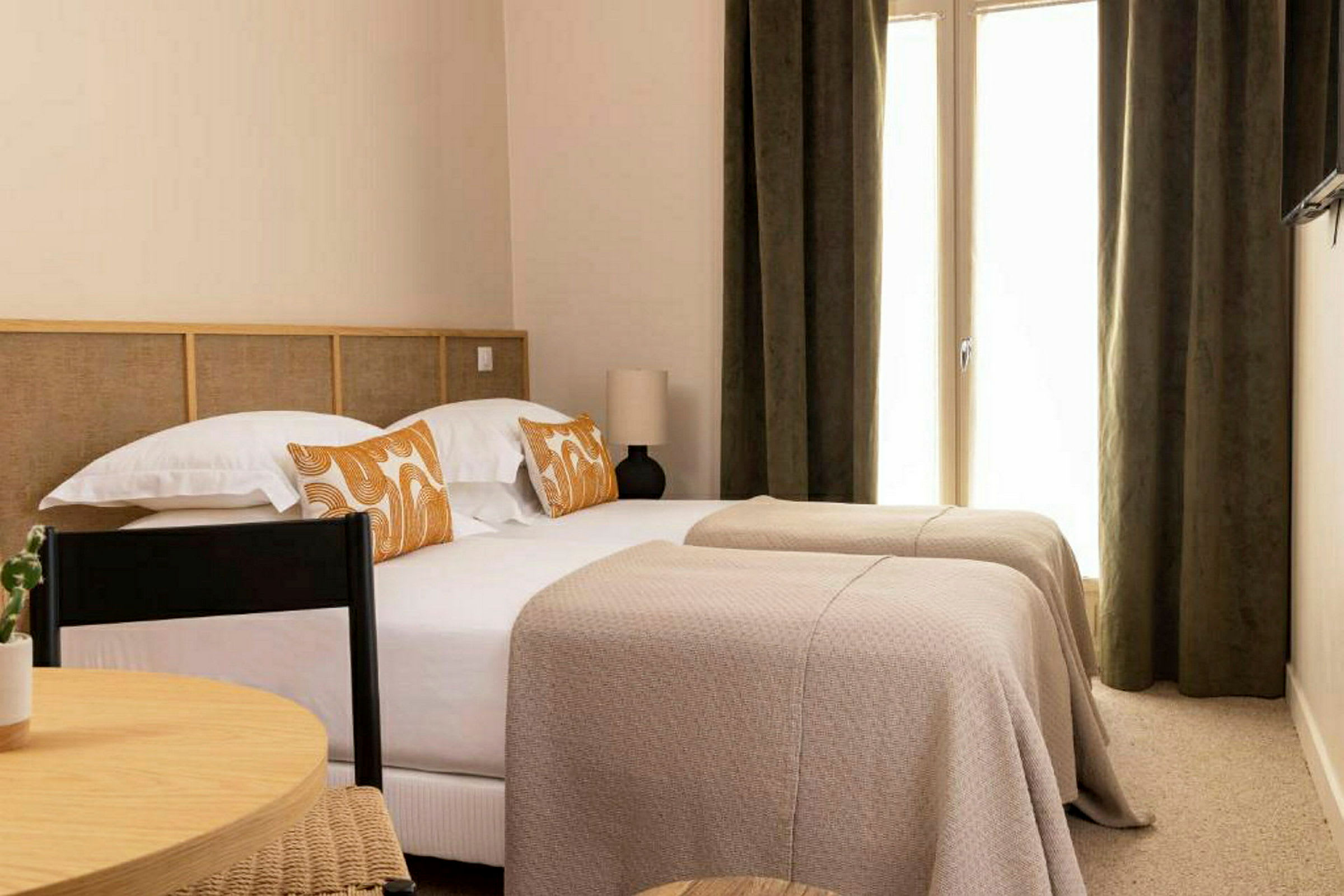 Ett hotellrum med två enkelsängar, täckta av beigea överkast och dekorerade med orange mönstrade kuddar. (AI-genererad text)