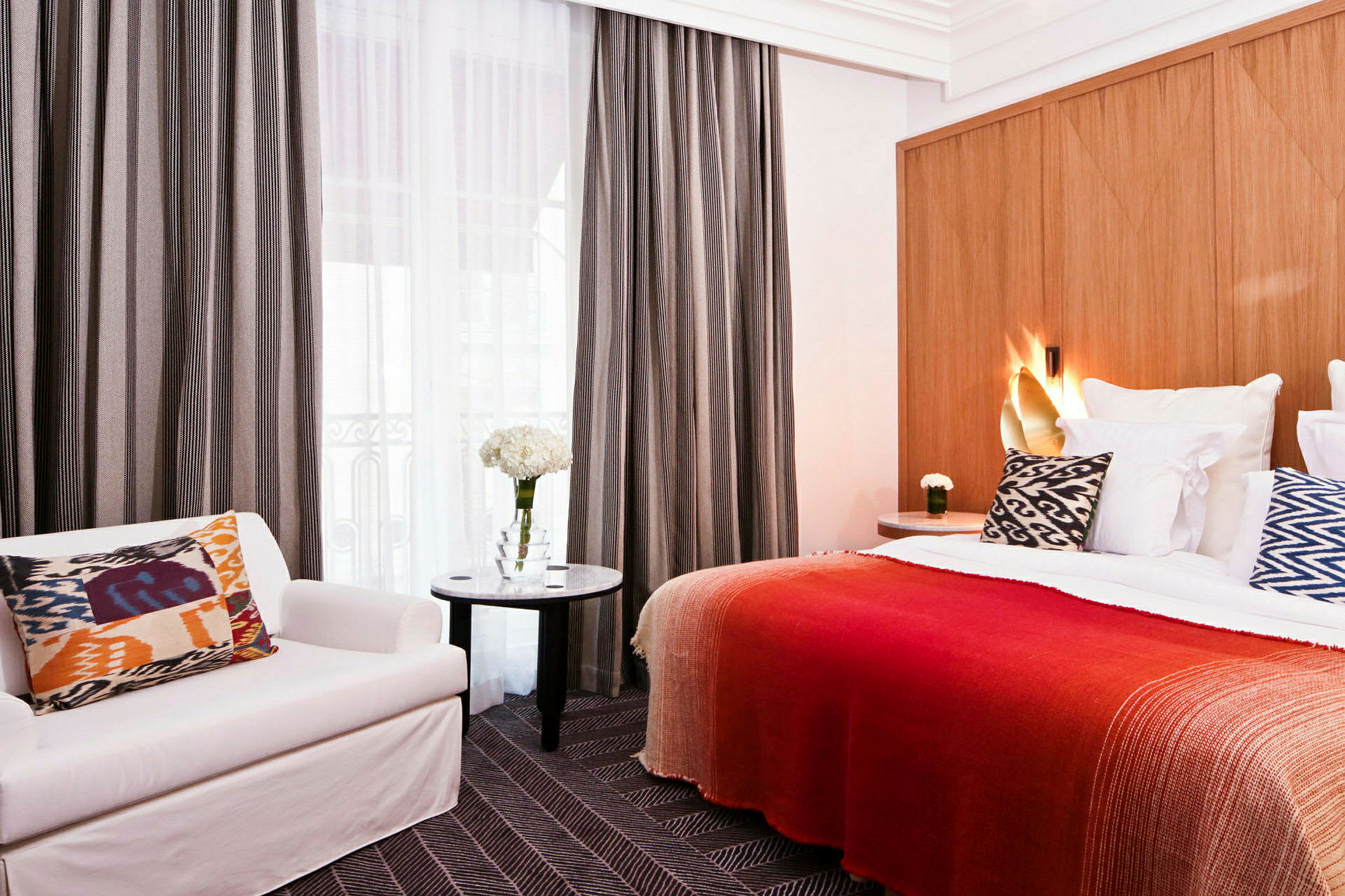 Hotellrum med säng, kudde med mönster, röd filt, stol och bord med blommor. (AI-genererad text)