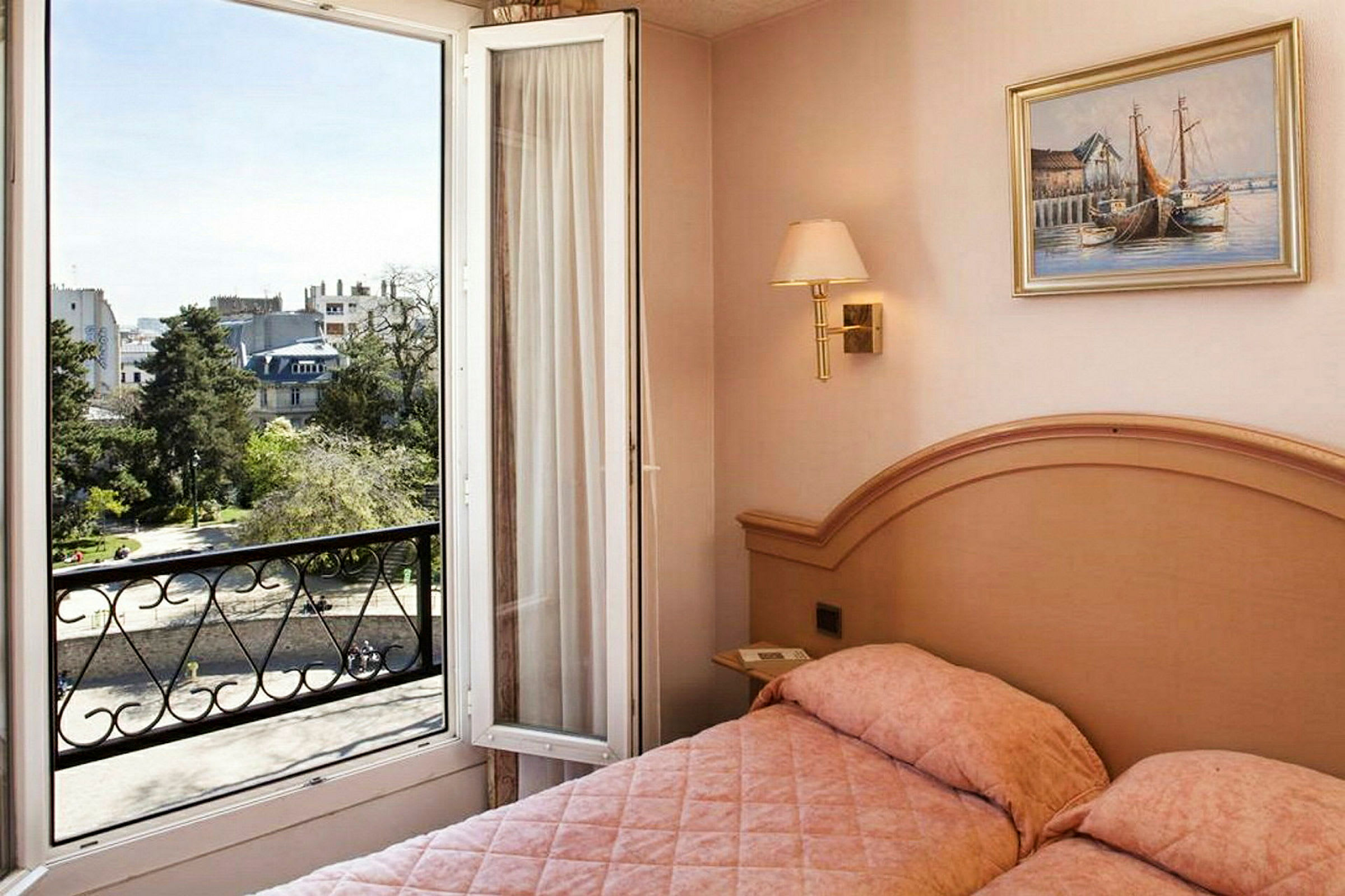 Ett hotellrum med en säng och en öppen balkongdörr som visar utsikt över en stad och grönområde. (AI-genererad text)