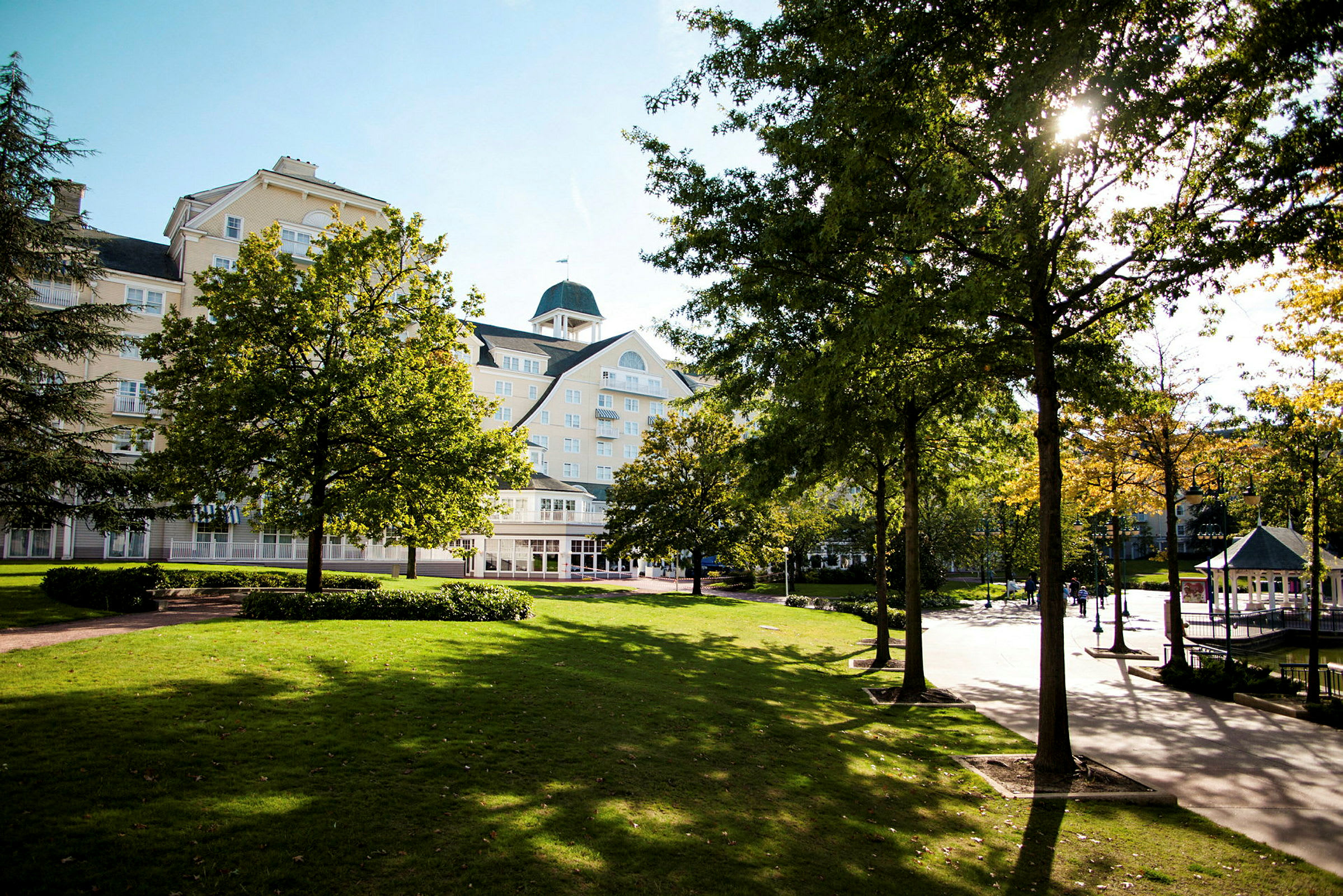En park med gröna träd och gräsmatta framför en större byggnad under en solig dag. (AI-genererad text)