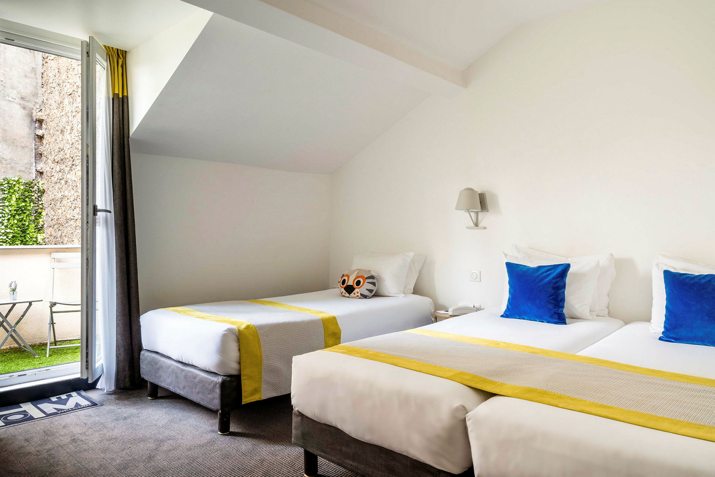 Ett hotellrum med två enkelsängar, dekorerade med blå kuddar och gulrandiga överkast, samt en öppning till en liten uteplats. (AI-genererad text)