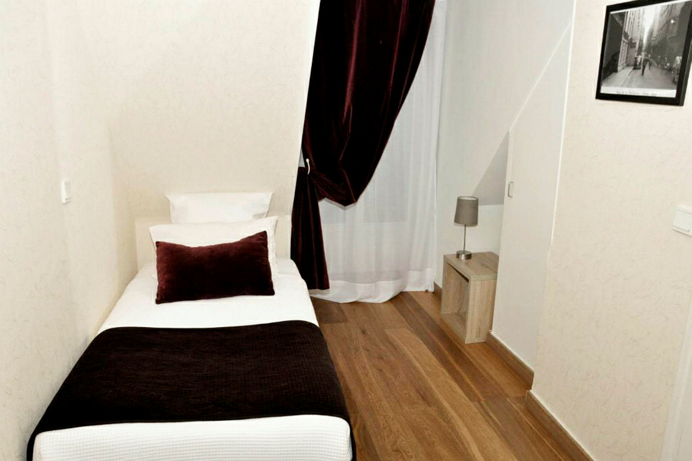 Ett hotellrum med enkelsäng, nattduksbord och en lampa. Rummet har trälaminatgolv och en gardin framför ett fönster. (AI-genererad text)
