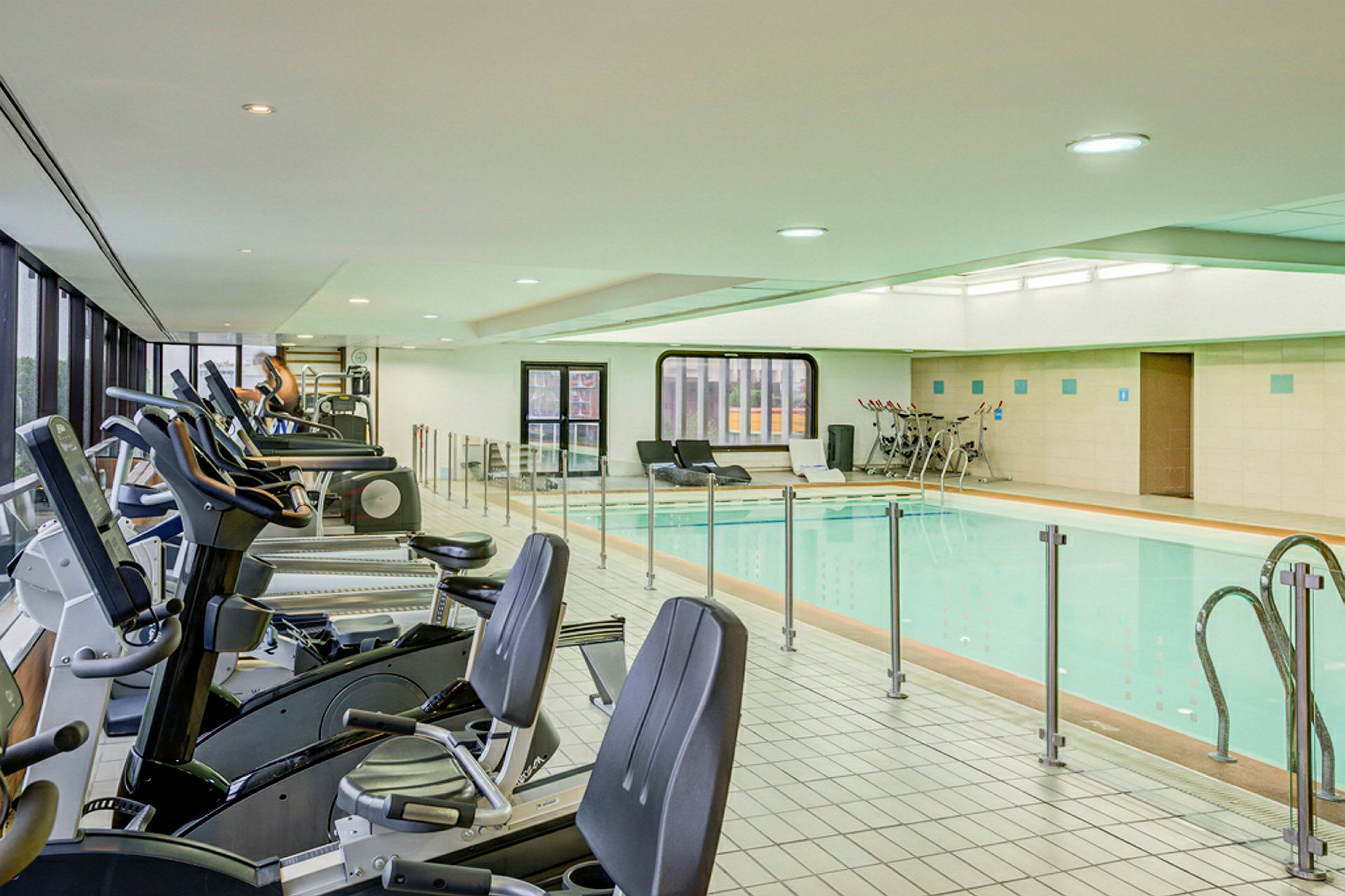 Ett gym med träningsmaskiner samt en bred simbassäng i en ljus inomhusmiljö. (AI-genererad text)