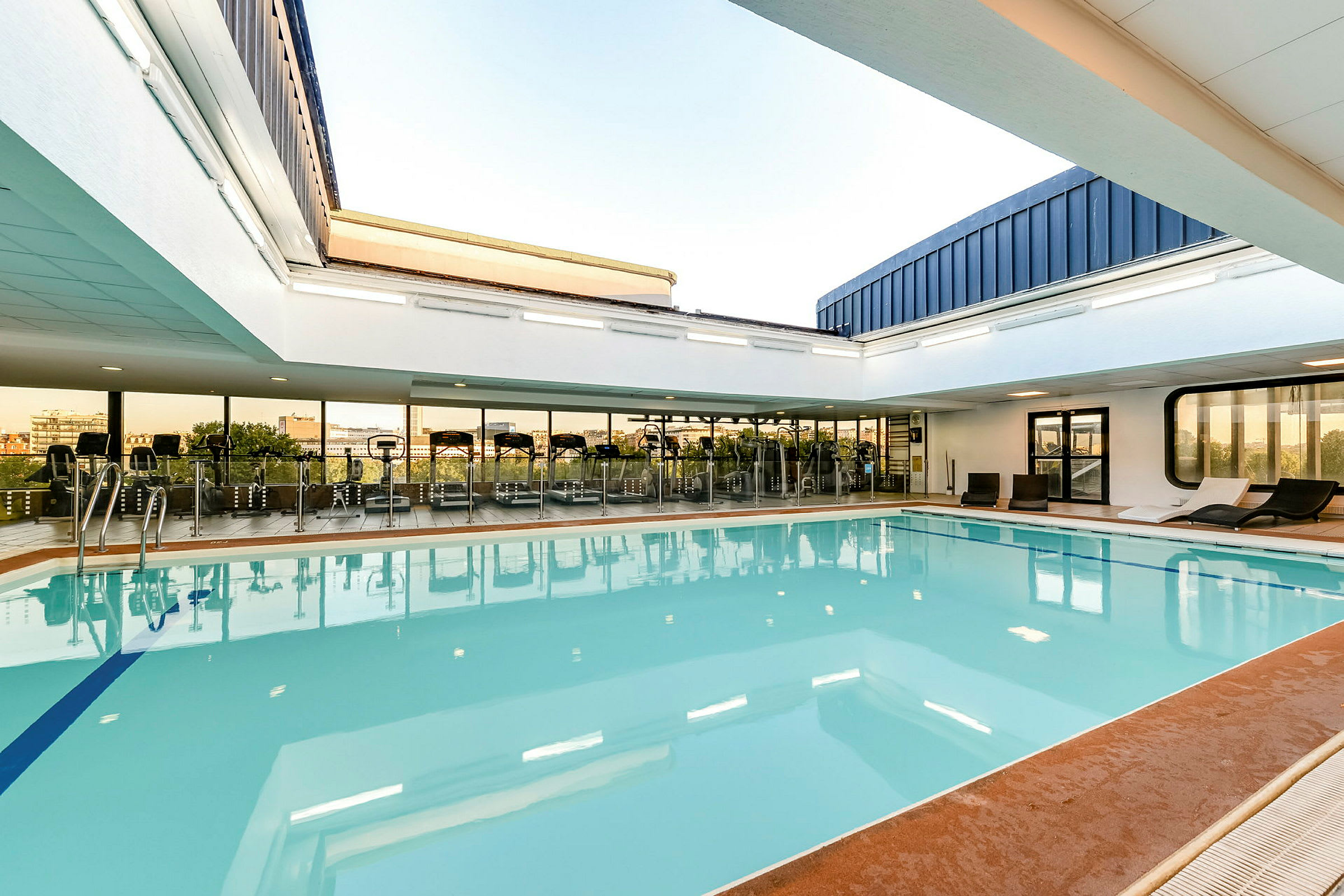 En inomhuspool med omgivande solsängar och stora fönster mot ett gym. (AI-genererad text)