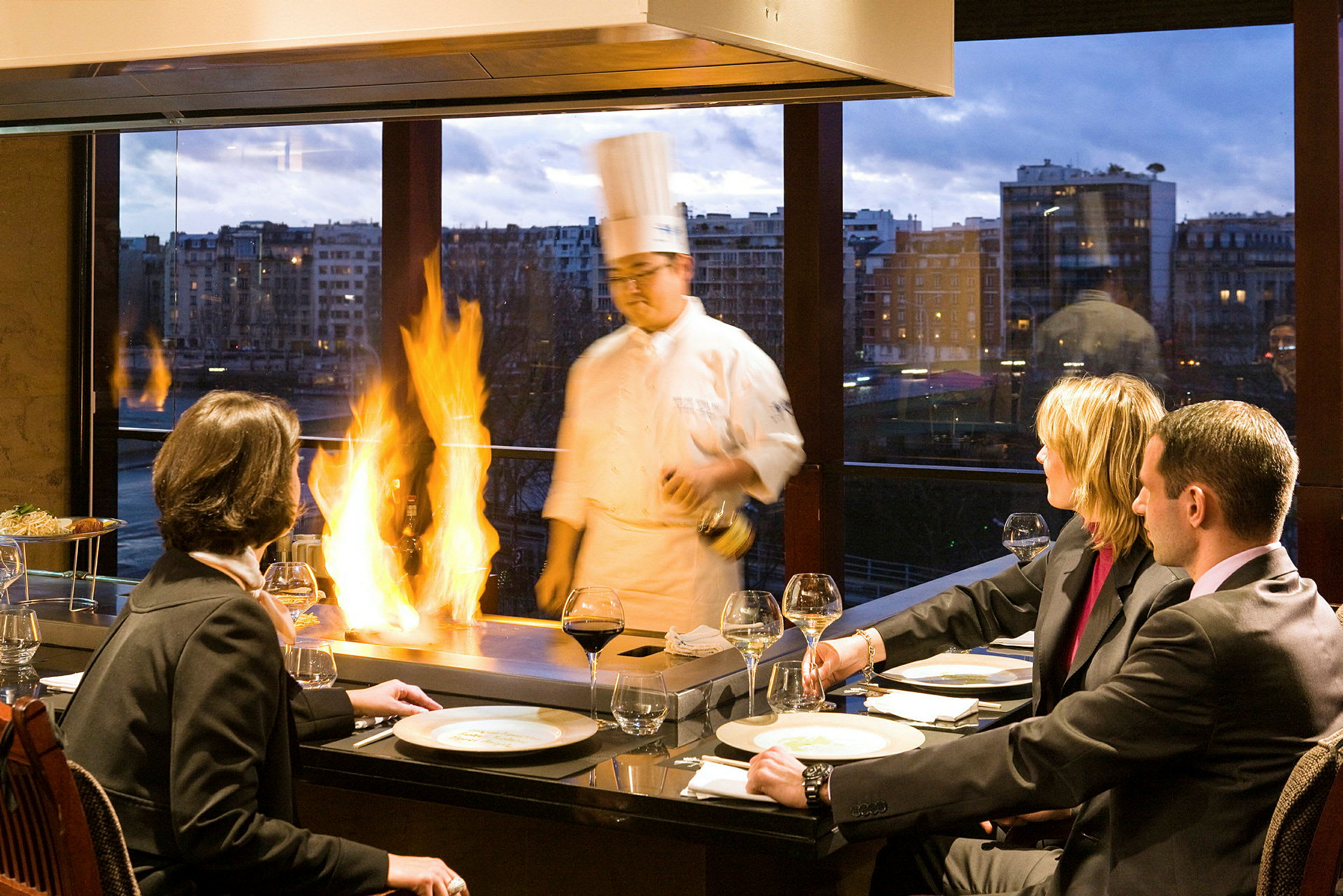 En grupp människor sitter vid ett bord och avnjuter en middag medan kocken tillagar mat med flammaffekt i en restaurang med stora fönster och kvällsutsikt. (AI-genererad text)