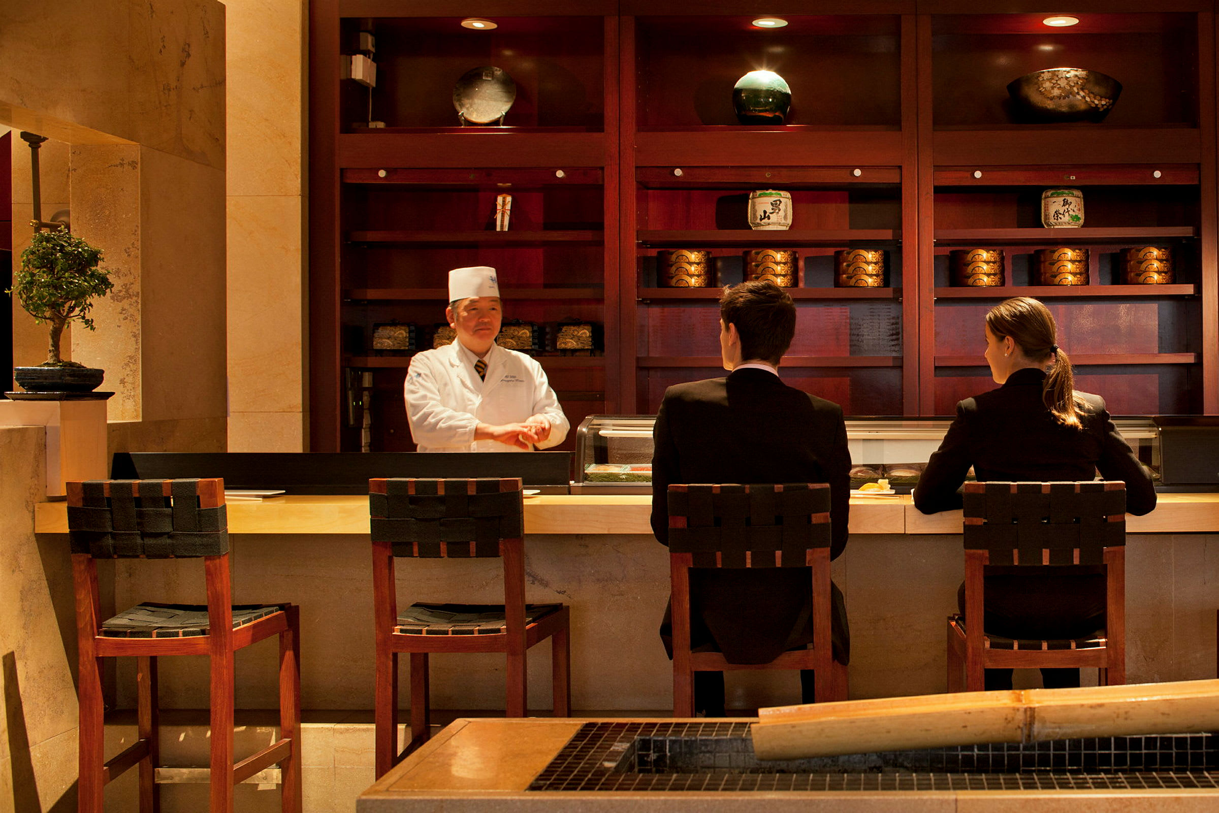 Två personer sitter vid en sushi-bar med en kock bakom disken. (AI-genererad text)