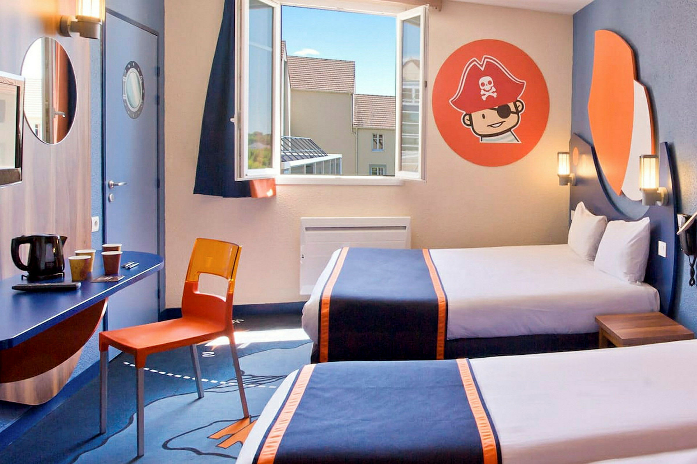 Ett hotellrum med två sängar försedda med mörkblå överkast och orangea detaljer. Det finns ett fönster med öppen utsikt, en vägg med ett sjörövartema på dekorativa väggmålningar samt ett skrivbord med en orange stol och kaffekokare under en rund spegel. (AI-genererad text)
