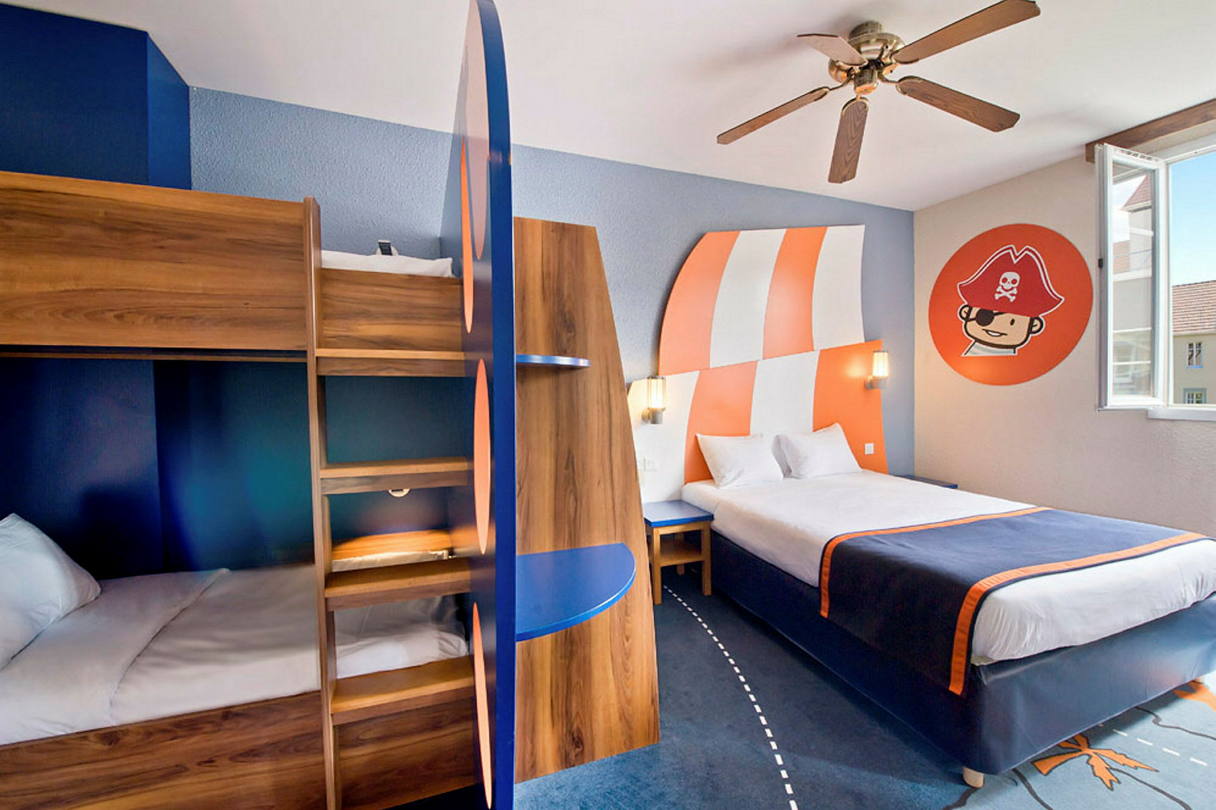 Ett hotellrum med ett våningssäng i trä, en dubbelsäng, dekor i blått och orange samt en väggmålning med piratmotiv. (AI-genererad text)