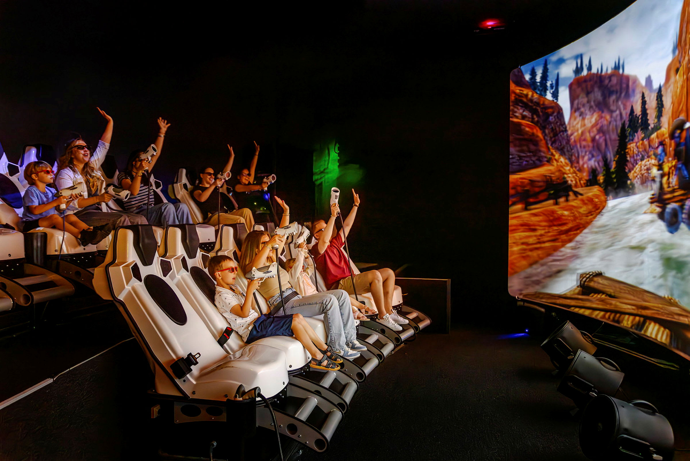 En grupp människor sitter i en interaktiv simulator med en stor skärm som visar en naturscen med berg och flod. (AI-genererad text)