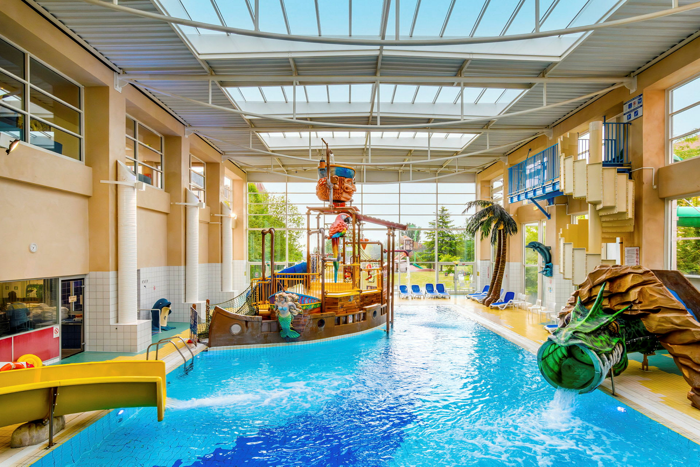 En inomhuspool med ett piratskepp och vattenrutschbanor, omgiven av solstolar och växter. (AI-genererad text)