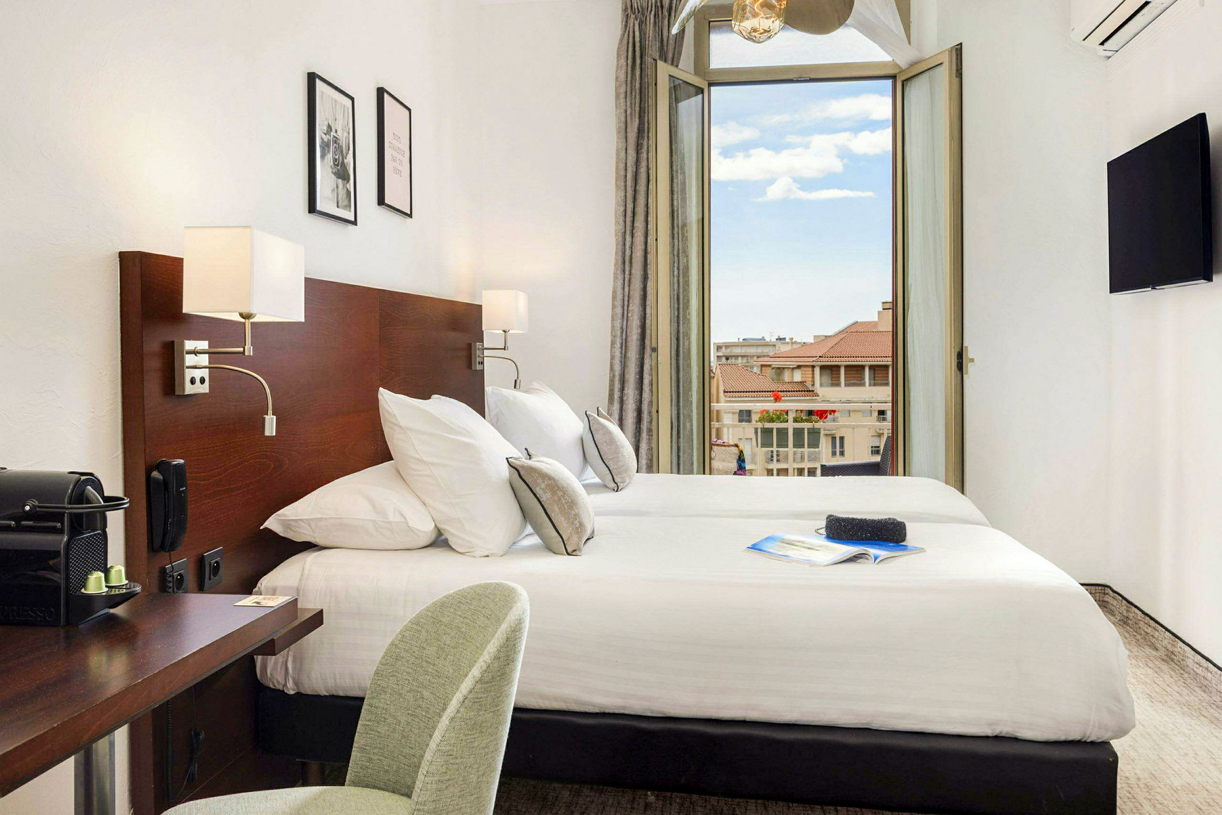 Ett hotellrum med dubbelsäng, skrivbord och en öppnad balkongdörr som visar utsikt över omgivande byggnader. (AI-genererad text)