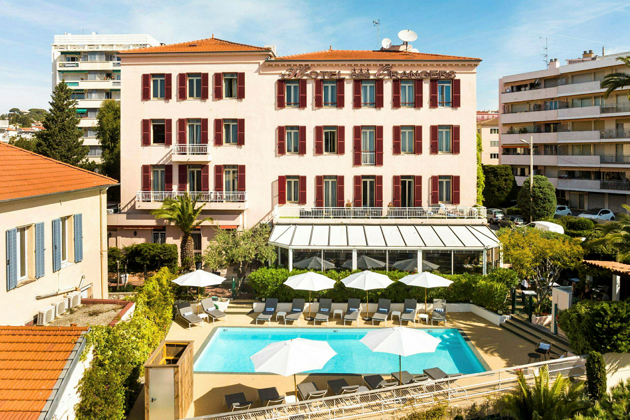 Ett hotell med rosa fasad och röda fönsterluckor, framför en pool med solstolar och vita parasoller. (AI-genererad text)