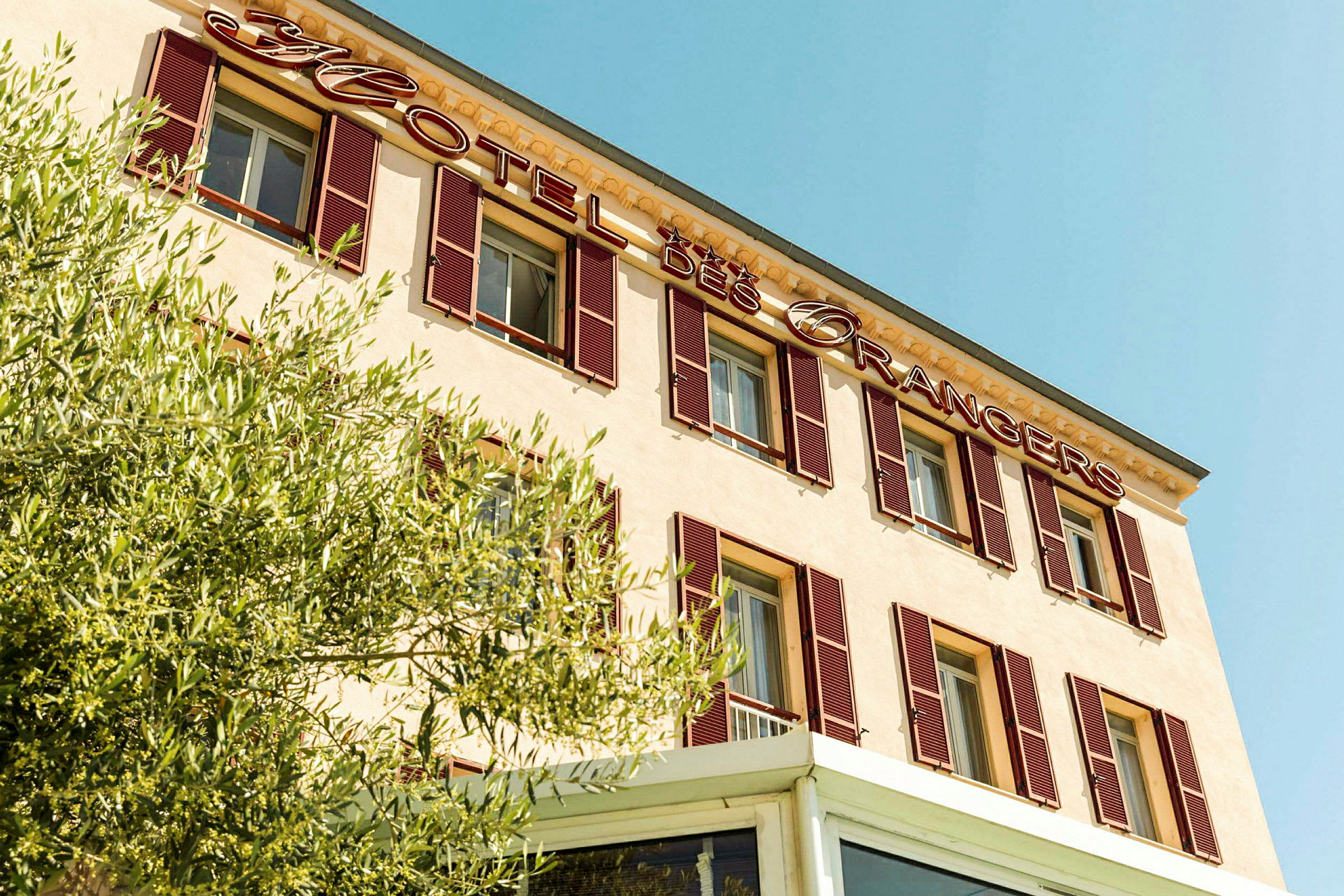 Fasad av hotellbyggnad med skylten 'Hotel des Orangers' och grönska i förgrunden. (AI-genererad text)