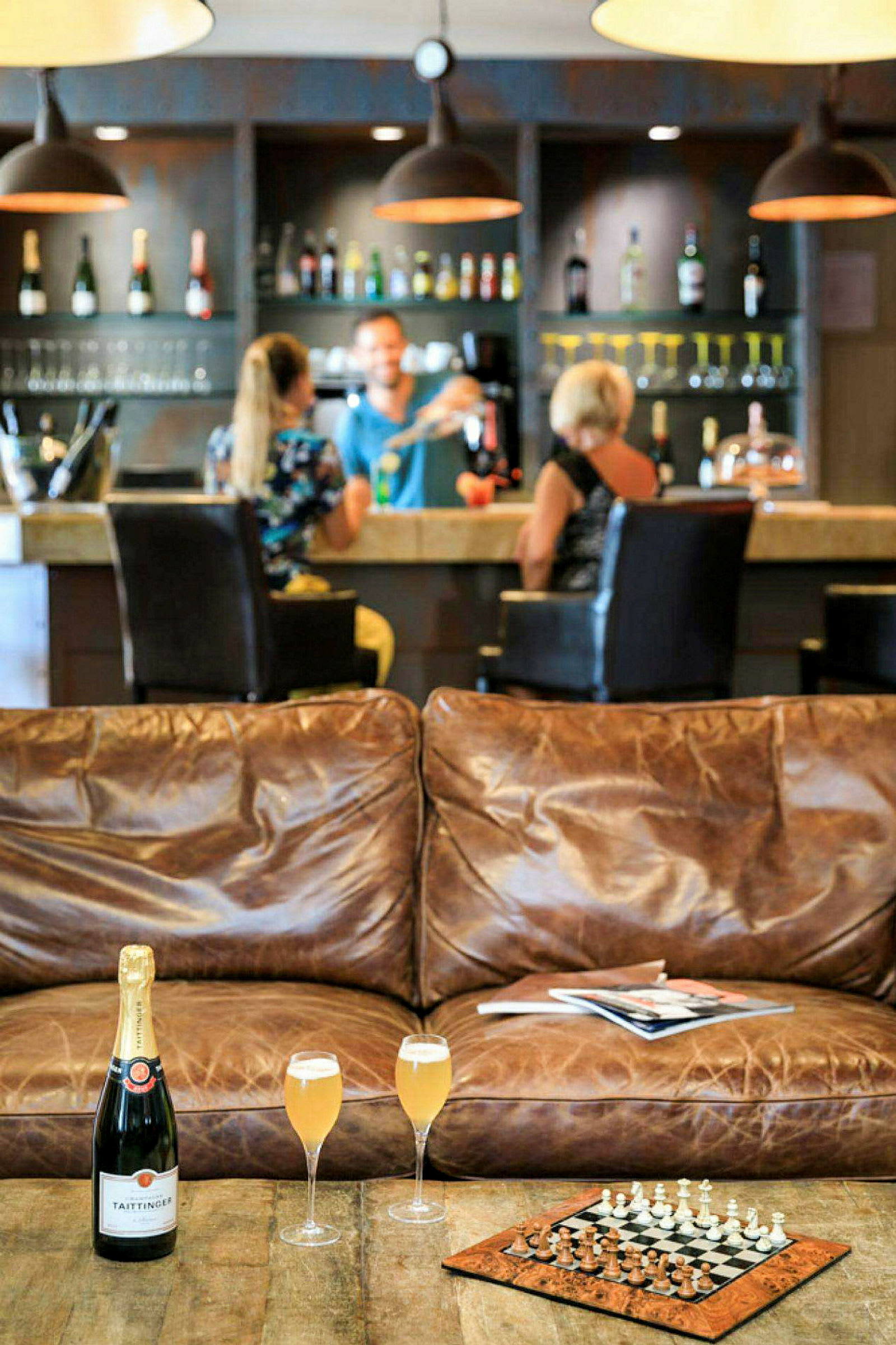 Interiörbild av en bar med dryckesflaskor i bakgrunden, en lädersoffa, ett schackspel och ett bord med champagneglas i förgrunden. (AI-genererad text)
