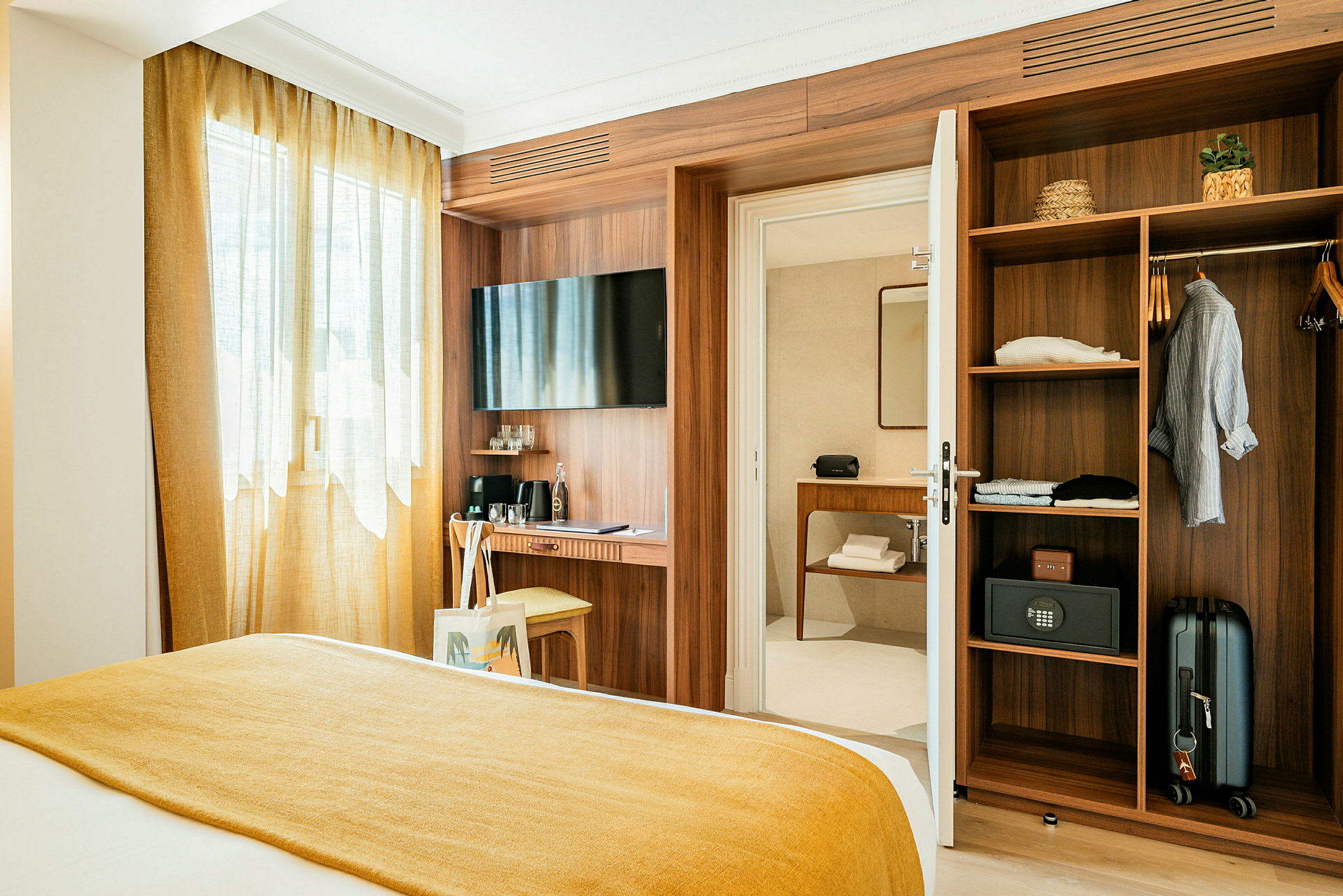 Ett hotellrum med en säng täckt av en gul filt. Till höger finns en öppen garderob med kläder och en resväska. En dörr leder till ett badrum. (AI-genererad text)