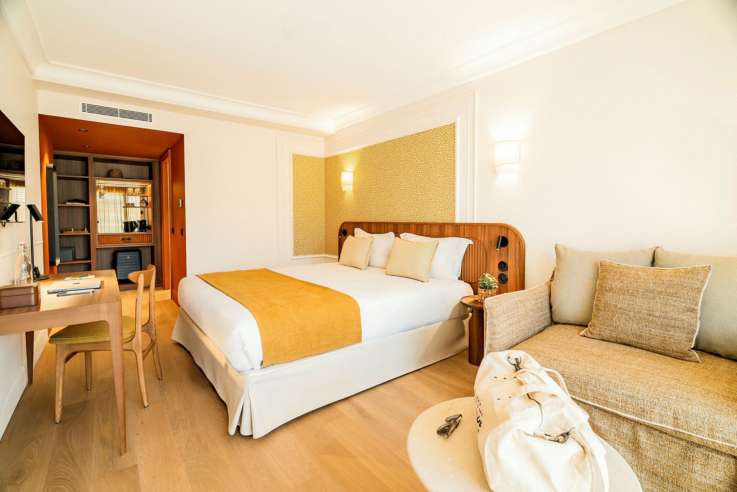 En hotellrumsinteriör med en dubbelsäng med gul överkast och kuddar, ett skrivbord med stol till vänster och en soffa med beige kuddar till höger. (AI-genererad text)