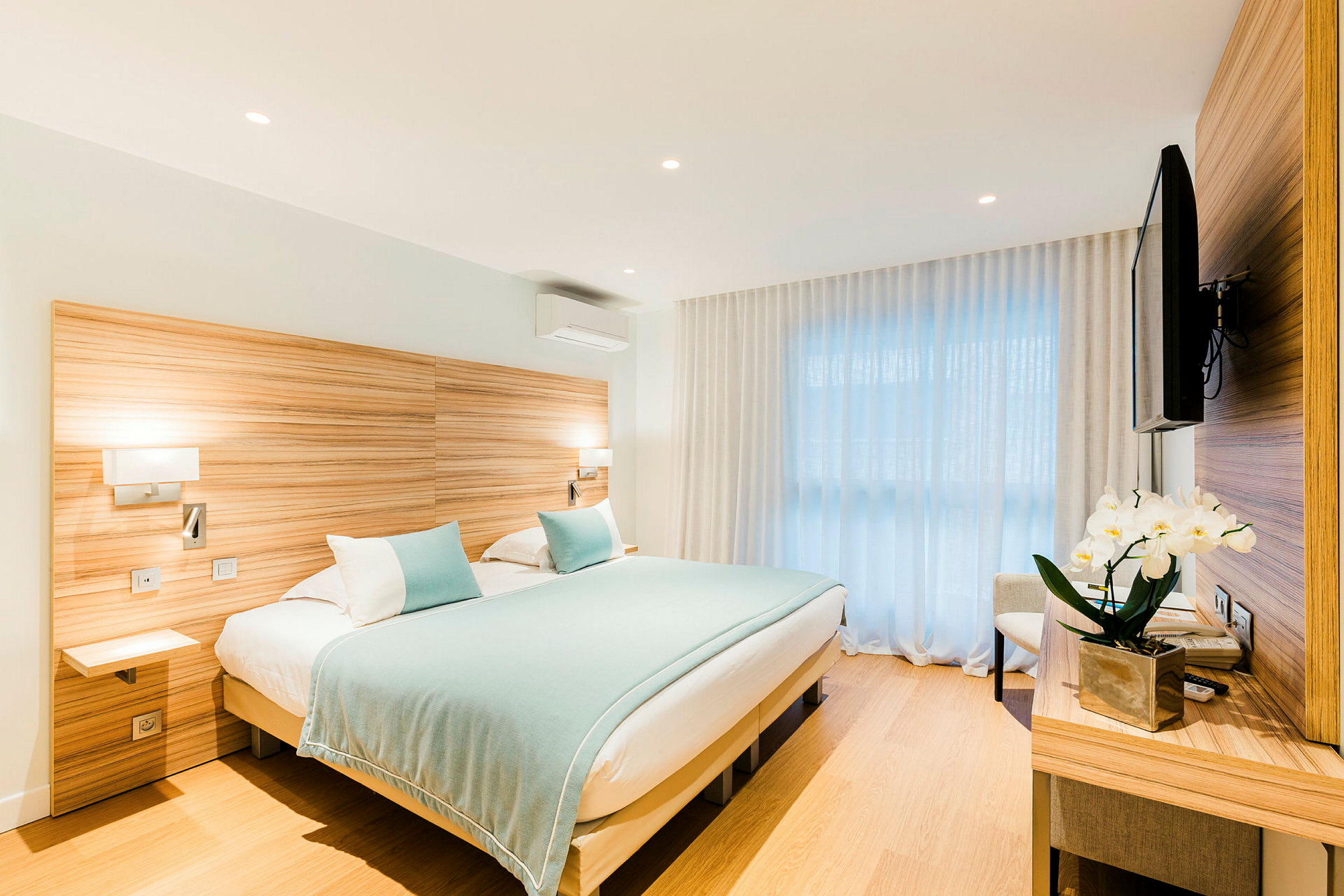 Ett hotellrum med en dubbelsäng klädd i ljusblå sängkläder och kuddar. Rummet har ljusa trämöbler, en väggmonterad TV och vita gardiner framför ett stort fönster. (AI-genererad text)