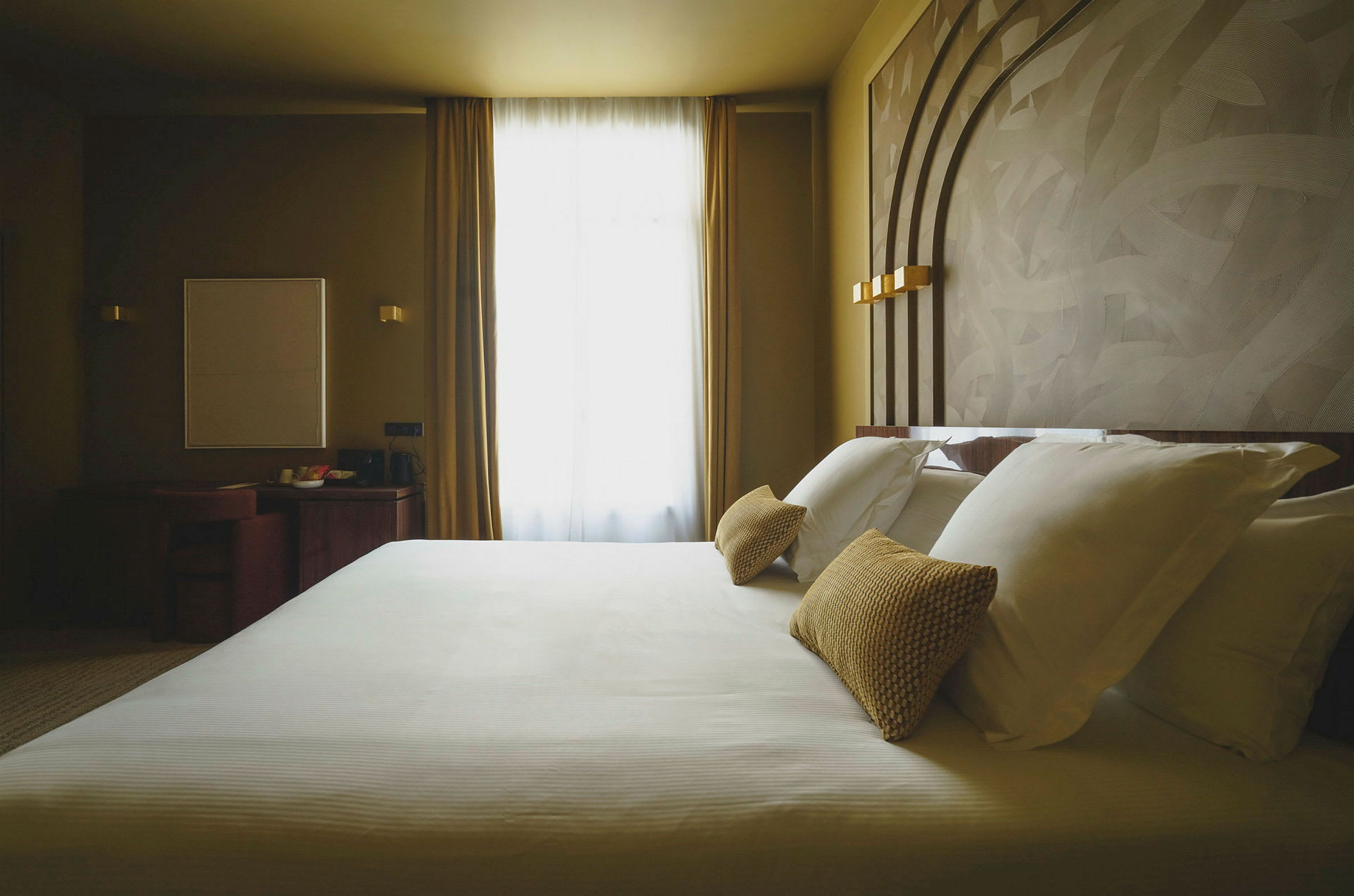 Hotellrum med en stor säng och dekorerande kuddar, fönster med gardiner i bakgrunden. (AI-genererad text)