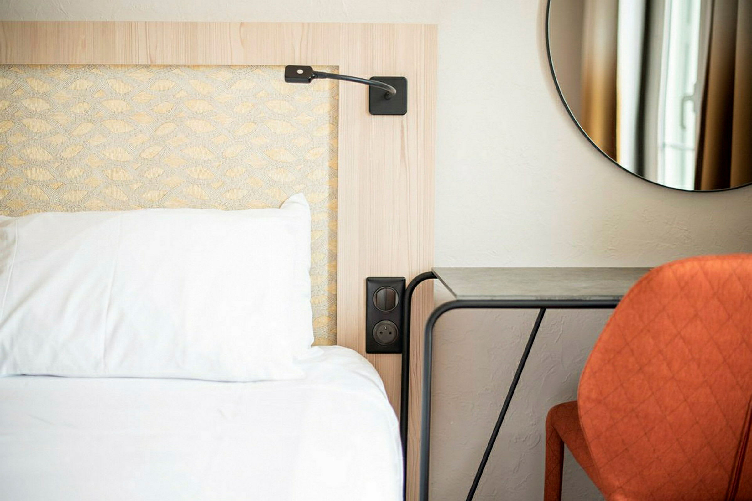 En hotellrumssäng med en vit kudde och ett nattduksbord bredvid, samt en orange stol framför ett bord och en rund spegel på väggen. (AI-genererad text)