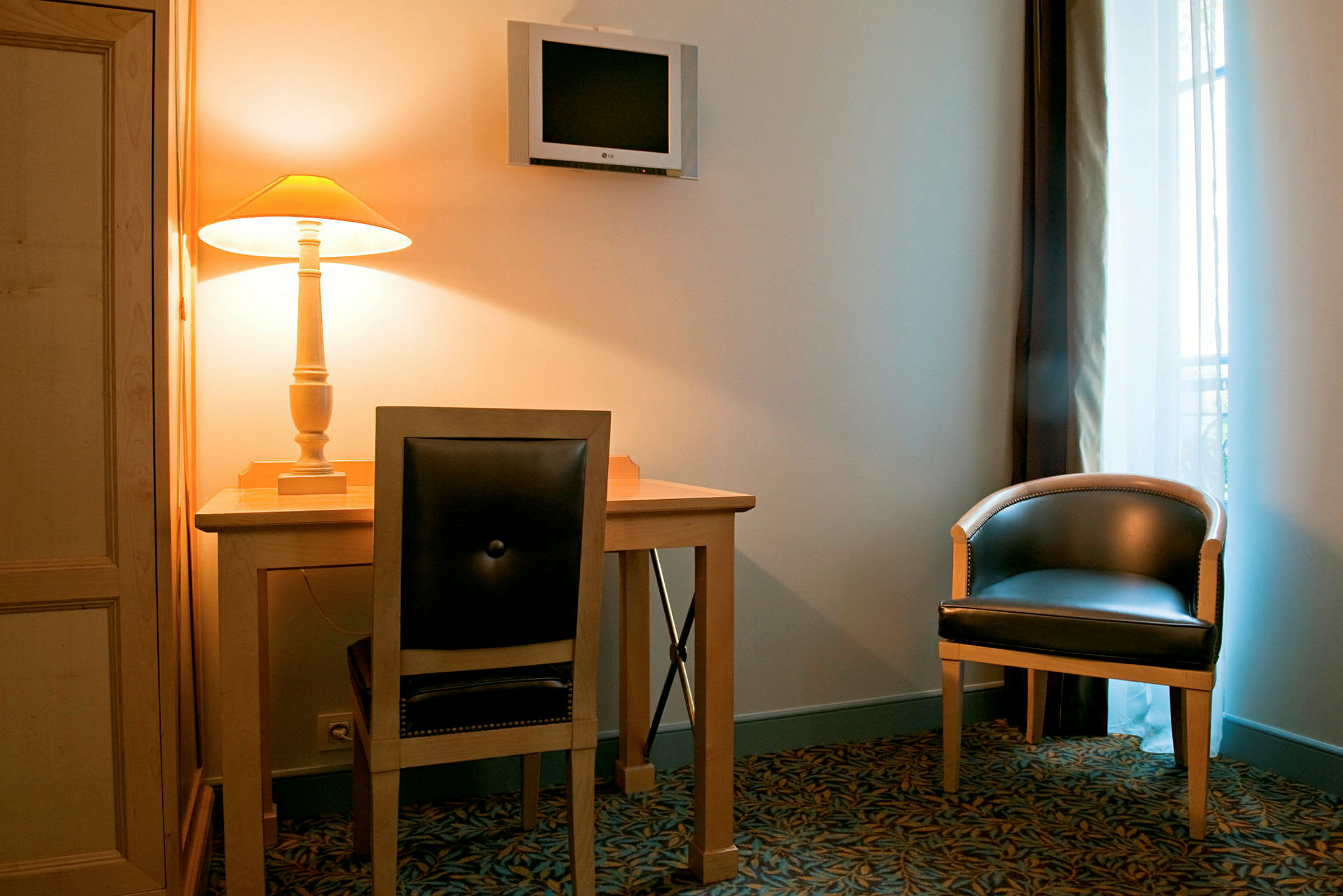 En inredning med ett skrivbord och stol, en väggfast TV samt en fåtölj bredvid ett fönster. Det står en tänd bordslampa på skrivbordet. (AI-genererad text)