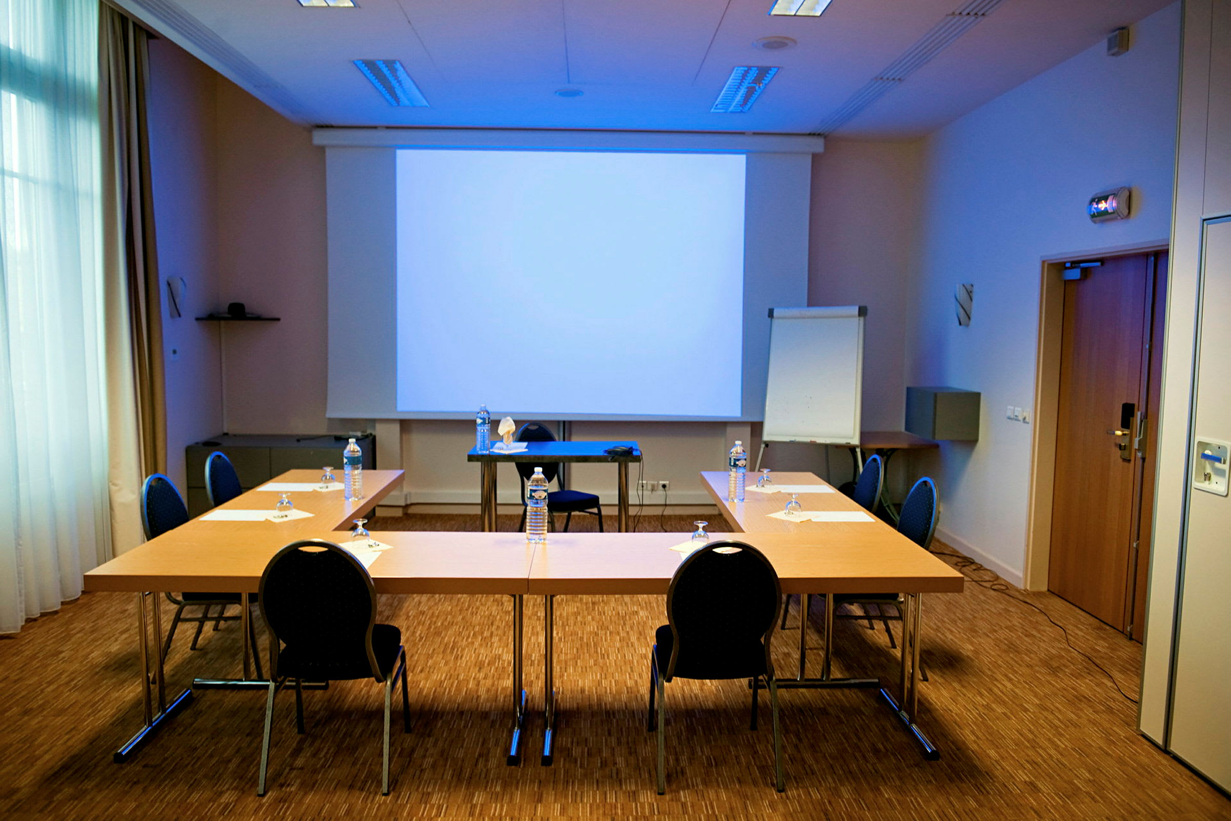 Ett konferensrum med ett uppsatt presentationsskärm, bord arrangerade i en u-form och stolar placerade runtom. (AI-genererad text)