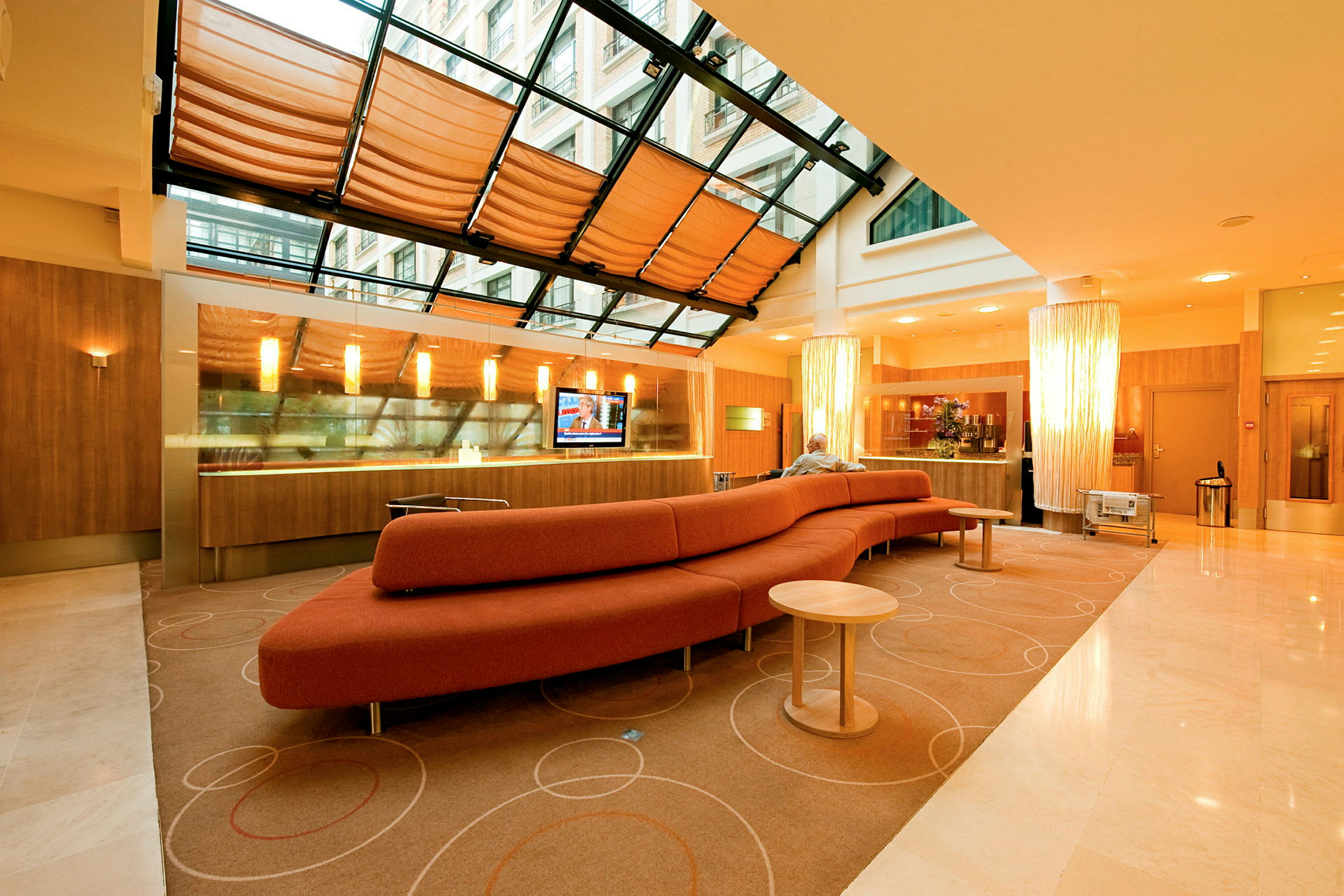 Ett modernt hotellobbyområde med en lång röd soffa, ett glastak med gardiner och ett receptionsområde i bakgrunden. (AI-genererad text)