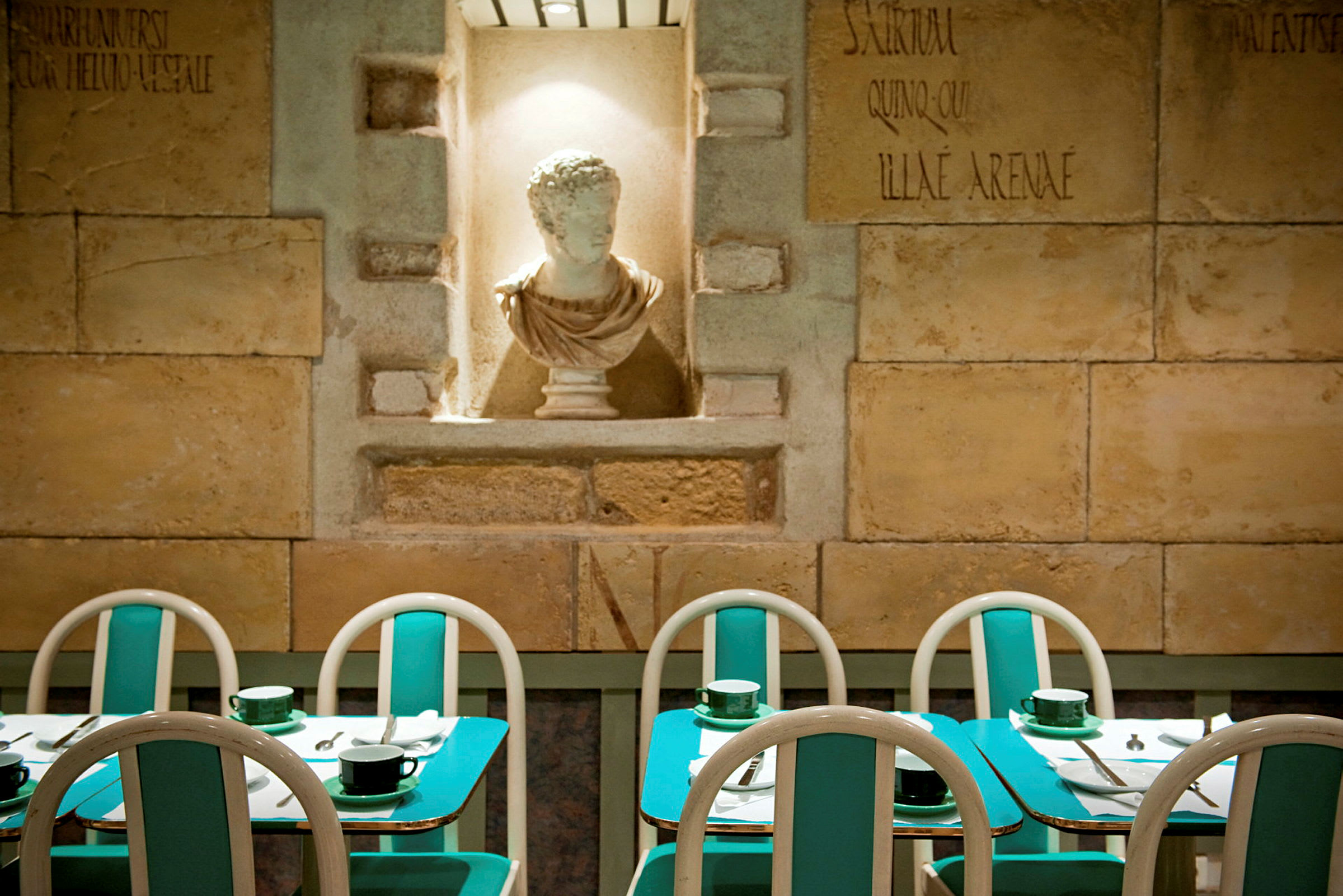 På bilden syns en restaurang med turkosa bord och stolar samt en vit skulptur i en nisch på väggen. (AI-genererad text)
