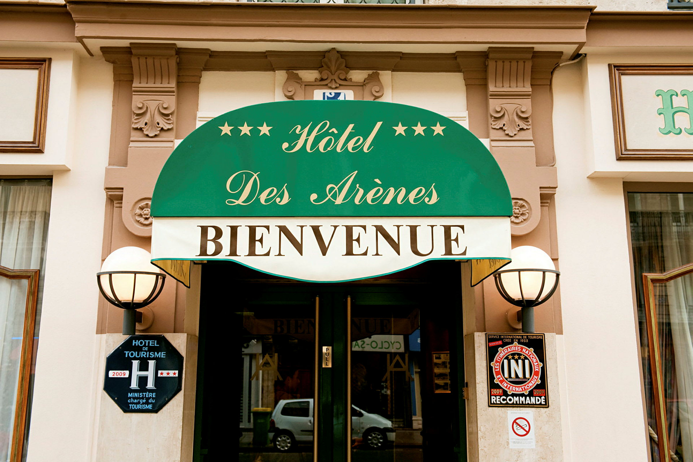 Entrén till Hôtel Des Arènes med grön markis och texten 'Bienvenue'. (AI-genererad text)